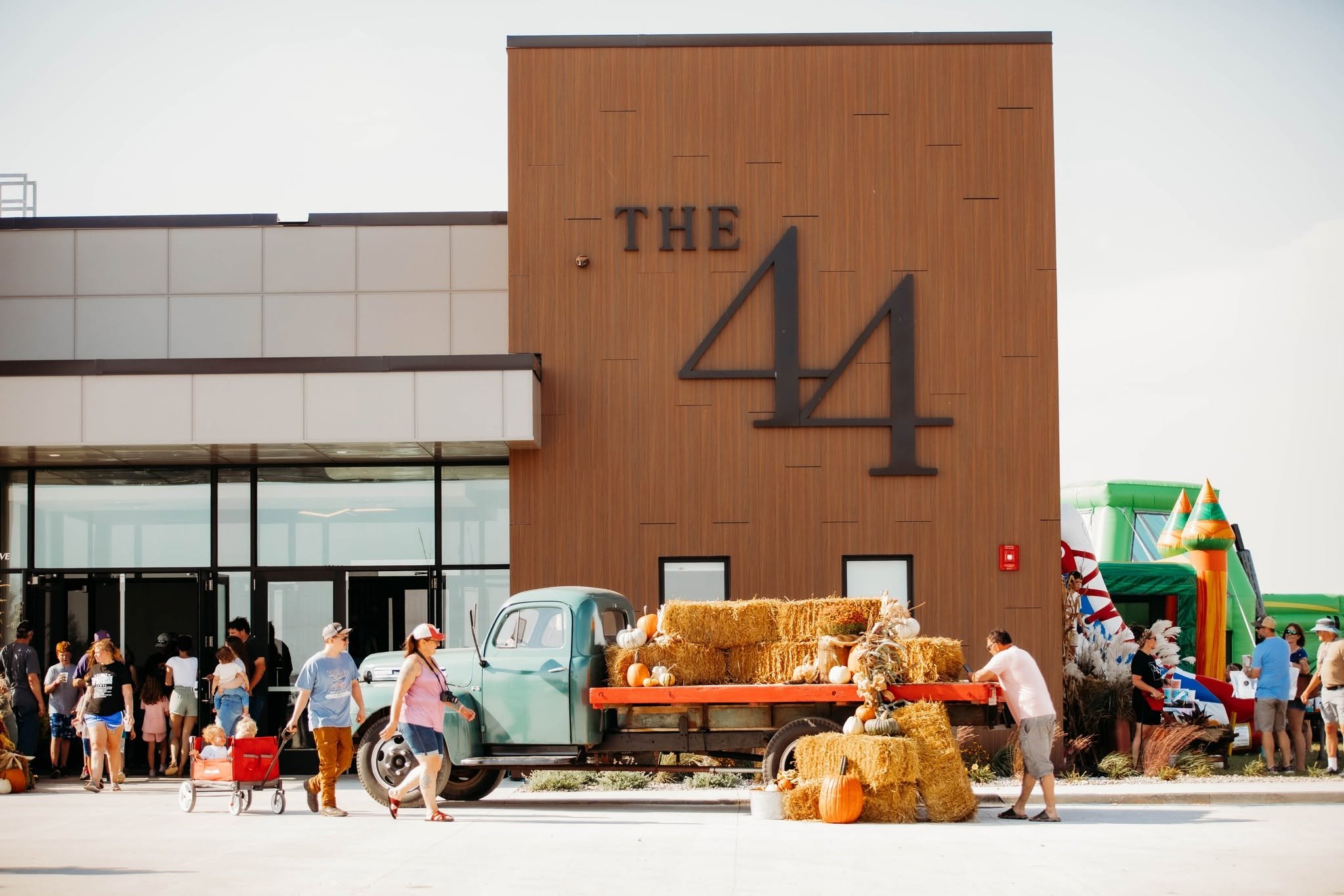 People gathered outside a building with a large wooden sign that reads 'The 44', decorated with hay bales and pumpkins for fall or Halloween. There is a vintage truck with more hay basks on it, and the scene suggests a festive outdoor event.