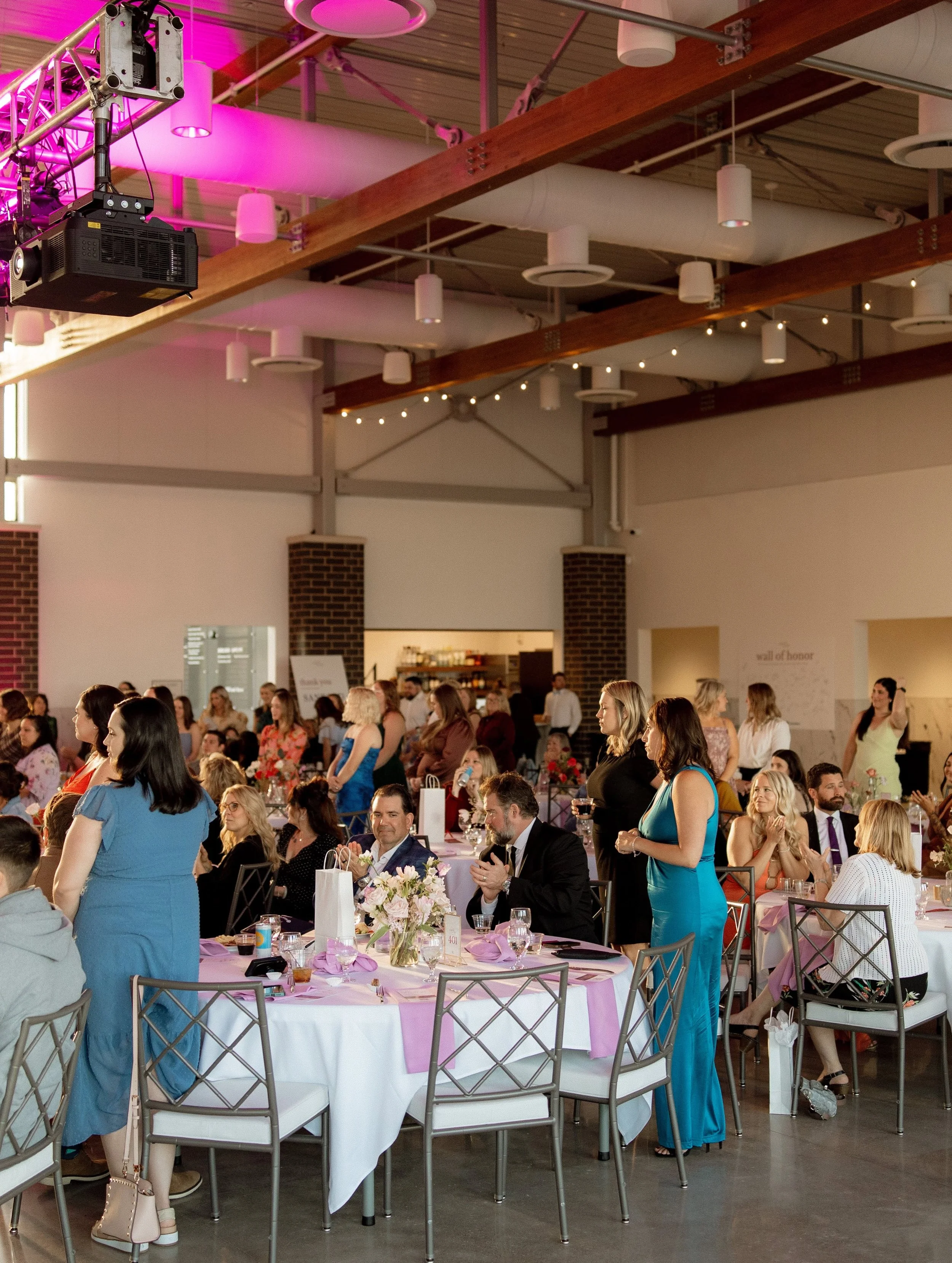 A large indoor event with many women seated and standing around decorated tables, some women are talking and some are looking around, with pink tablecloths, floral centerpieces, and various glasses and drinks, in a venue with wooden beams, pipe ceili