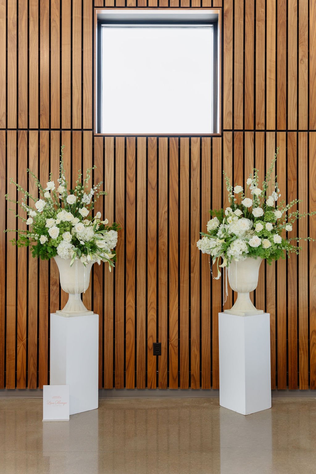 Two large white floral arrangements in urn vases on tall white pedestals in front of a wooden wall with vertical slats and a square window overhead.