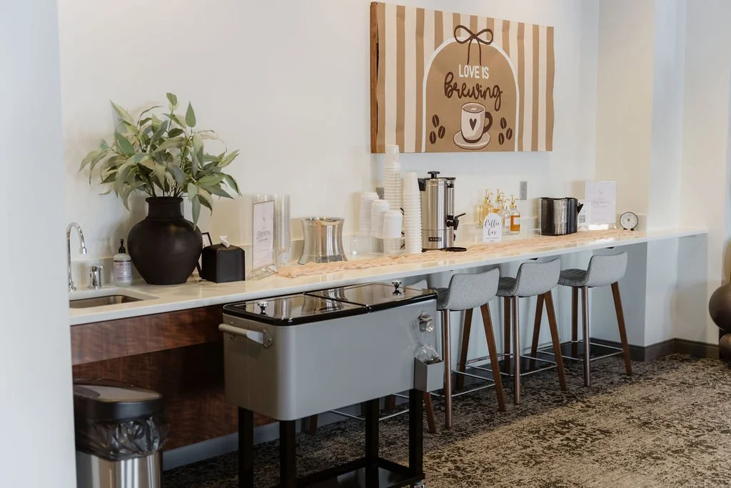 Coffee station with a large plant in a dark vase, coffee brewing equipment, disposable cups, and a sign that reads 'Love is Brewing' on a wooden wall art piece.
