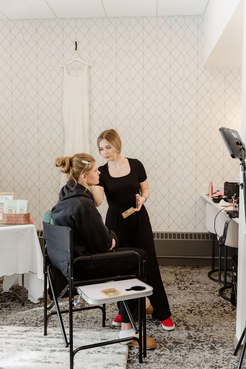 A makeup artist applying makeup to a seated woman in a black hoodie, in a room with a white patterned wall, a table with beauty products, and a mirror setup.