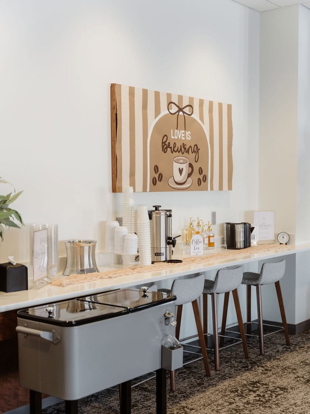 Coffee station with cups, coffee dispensers, and a sign saying 'Love is Brewing' in a cafe or break room.
