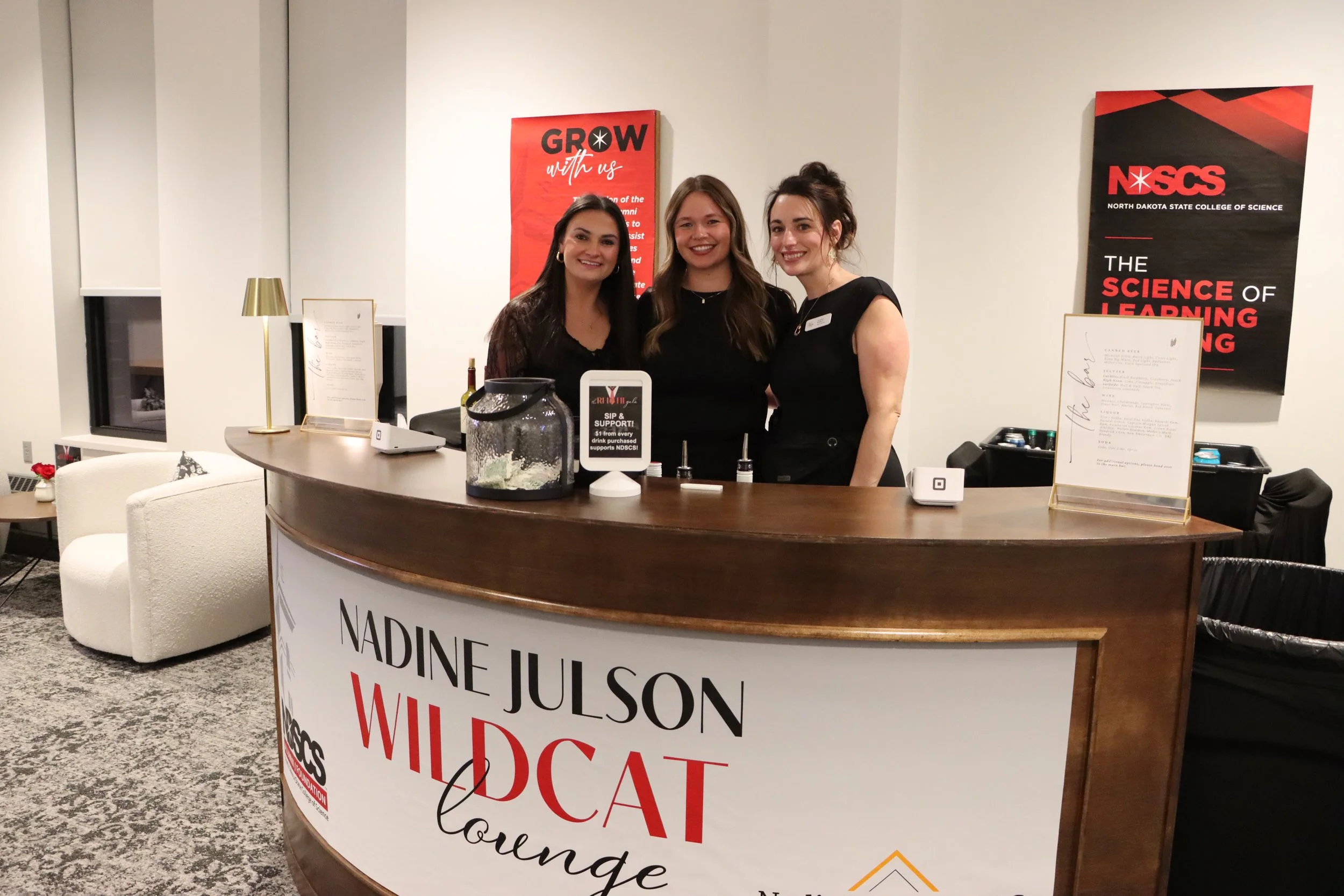 Three smiling women standing behind a curved reception desk with promotional signs and banners in the background, indicating a university event at North Dakota State College of Science.