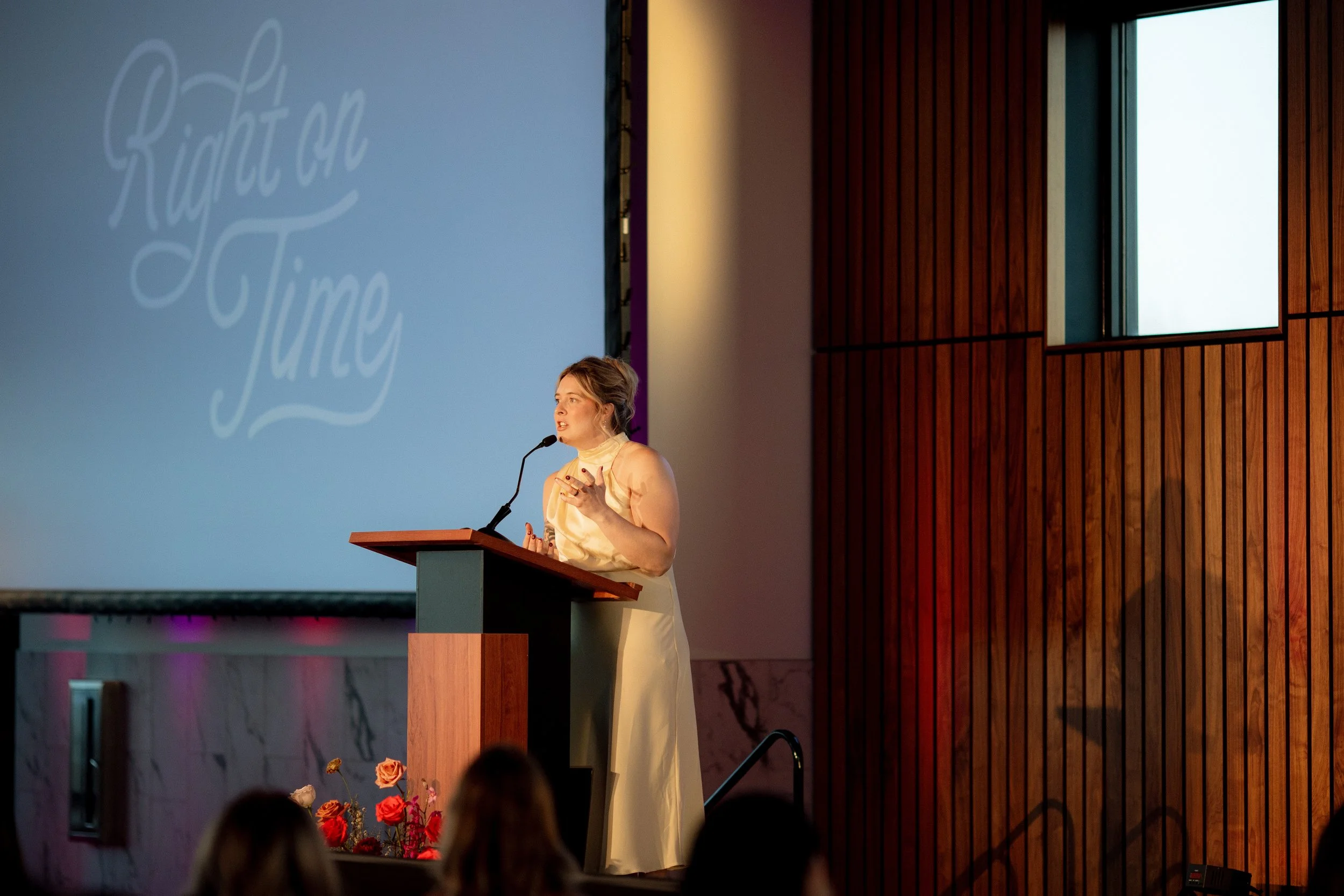 A woman in a yellow dress is standing at a podium, speaking into a microphone, with a large screen behind her displaying the words "Kapitolu Time" in script.