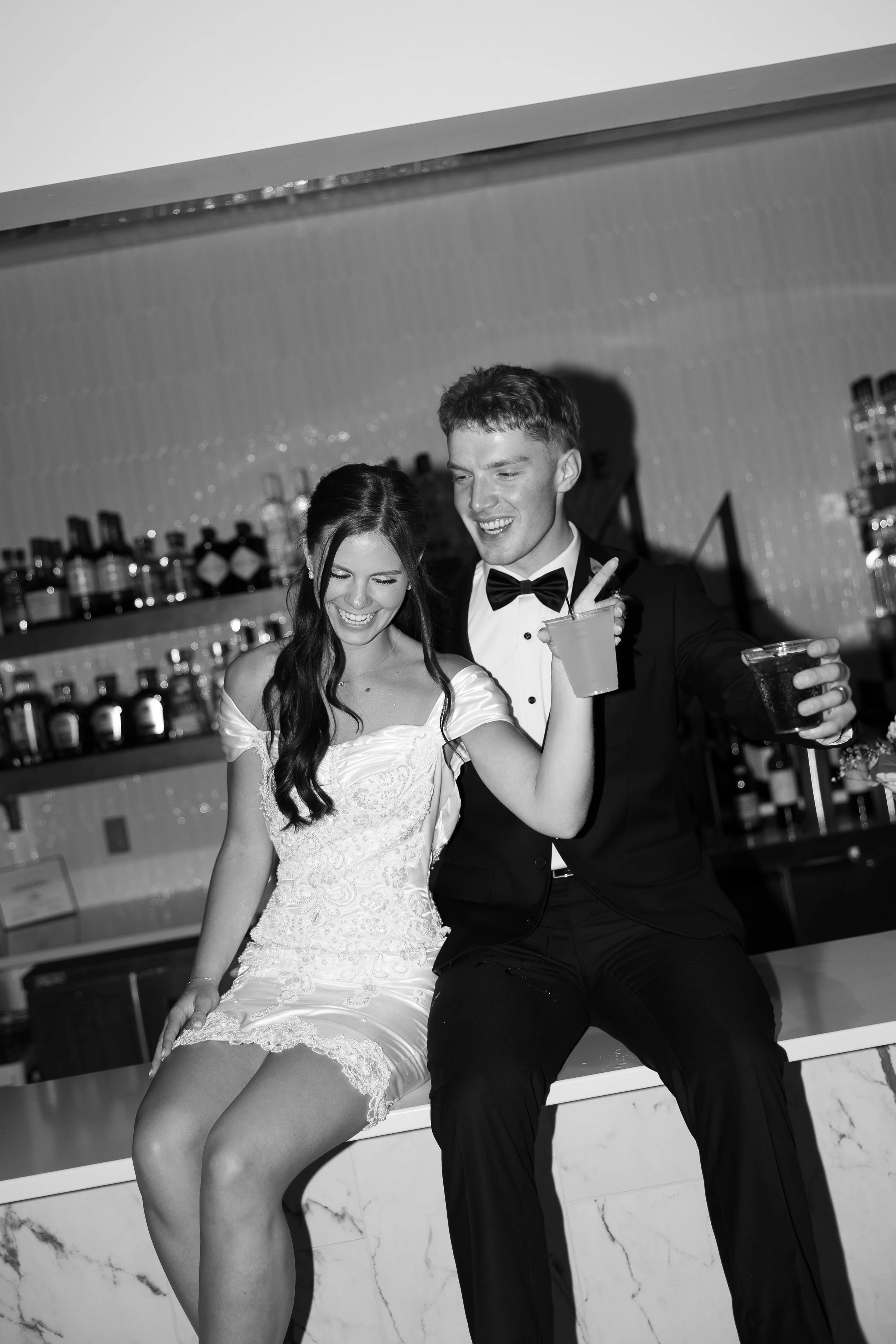A black-and-white photo of a newlywed couple laughing together while seated on the venue’s marble bar, each holding a drink. The bride wears a fitted lace wedding dress, and the groom wears a classic black tuxedo and bow tie.