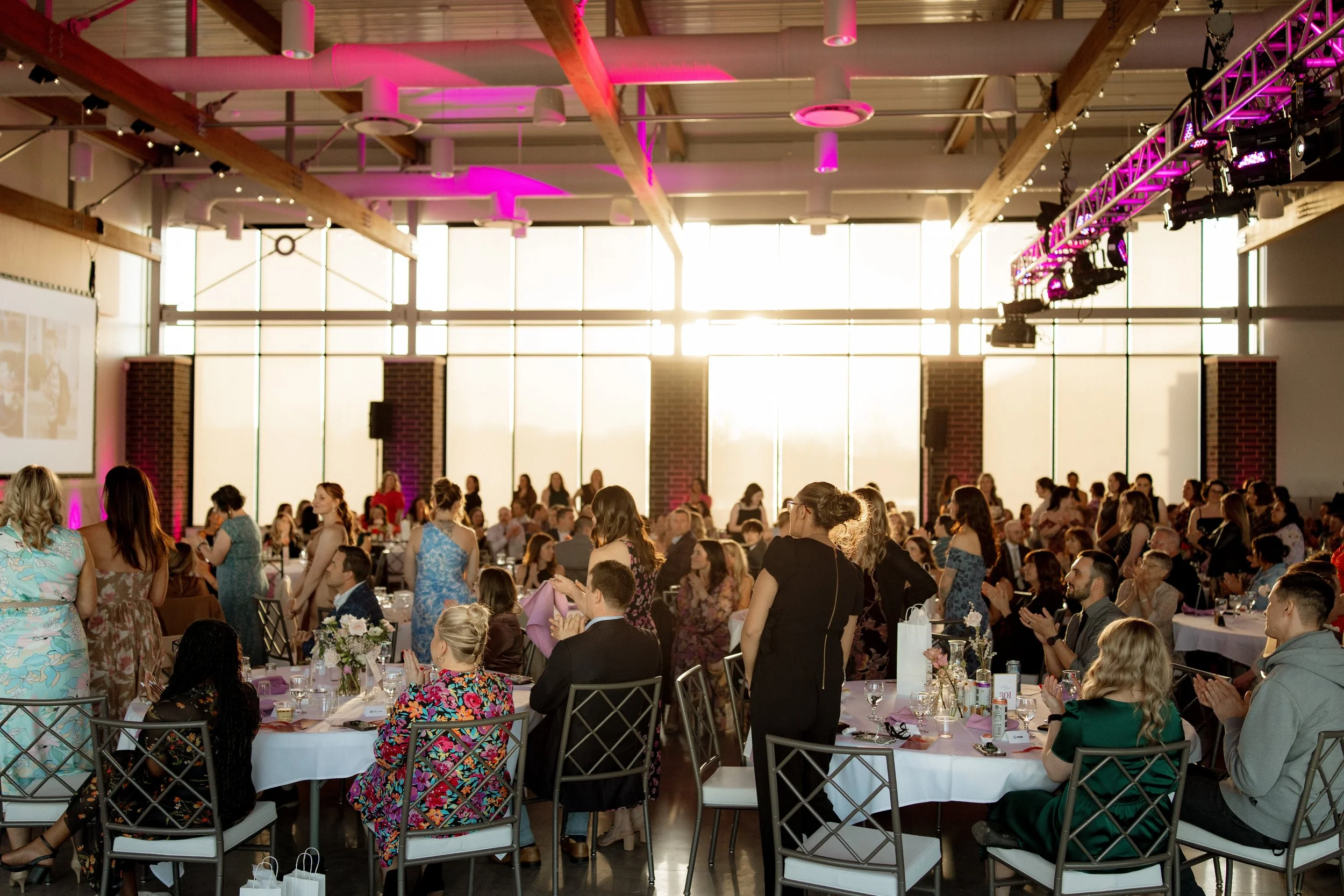People gathered at an indoor wedding reception, many wearing formal attire, sitting and standing around decorated tables, with large windows and stage lighting in the background.