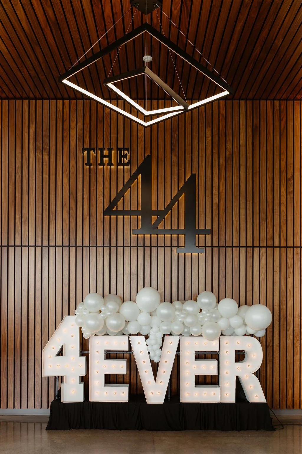Decorative wall with wooden paneling, featuring a large black number four in the center with the word "THE" above it, illuminated geometric ceiling lights, and a display of white balloons and marquee-letter lights spelling "FOREVER" at the bottom.