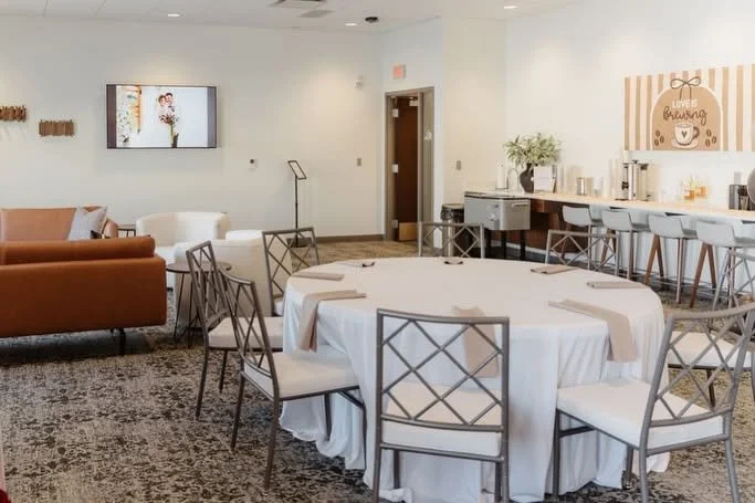 Interior of a modern event room with a large round dining table covered with a white tablecloth, surrounded by matching chairs, a bar area with white barstools, and a wall decorated with a sign that reads 'Gather, Breathe, Celebrate' with a coffee cu