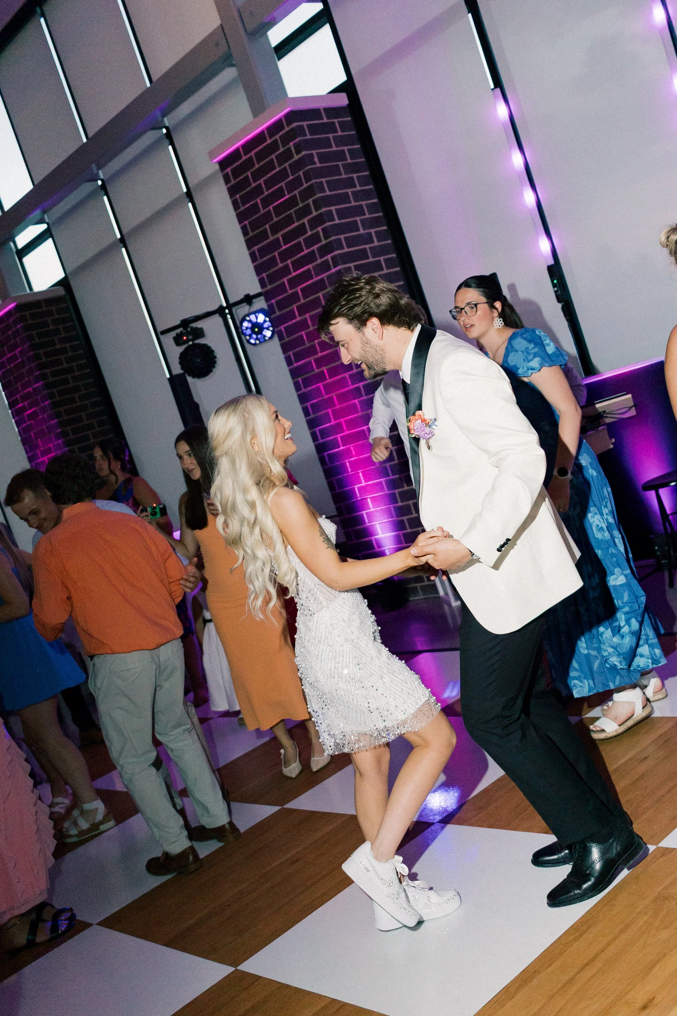 A newlywed couple smiles and dances together on our maple-and-white checkered dance floor, surrounded by guests and vibrant purple reception lighting from Harmon entertainment.