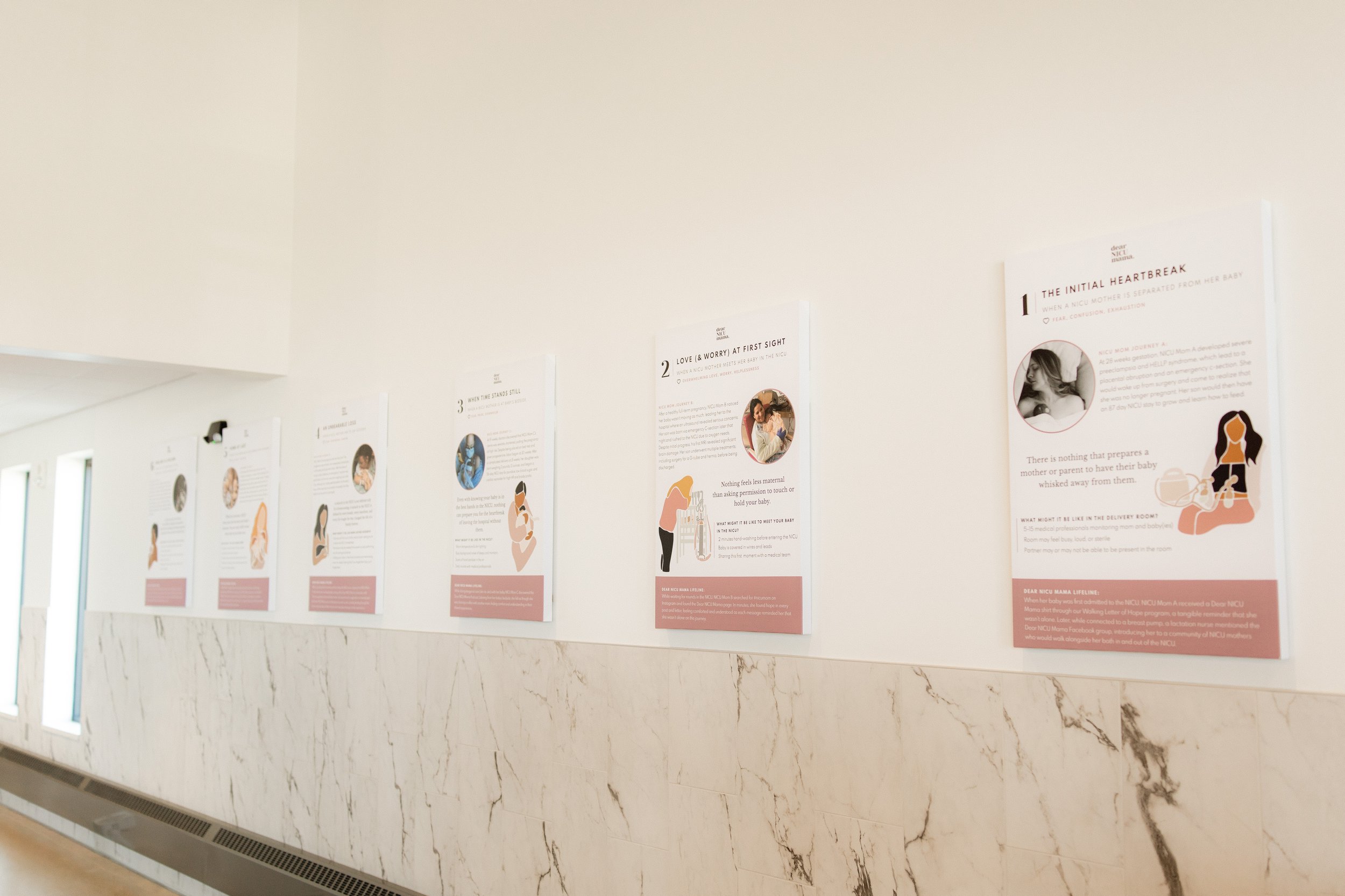 A hallway with white walls decorated with informational posters. The posters feature illustrations and text about NICU experiences and maternal topics, spaced evenly along the wall. The floor is beige with marble accents, and there are windows allowi