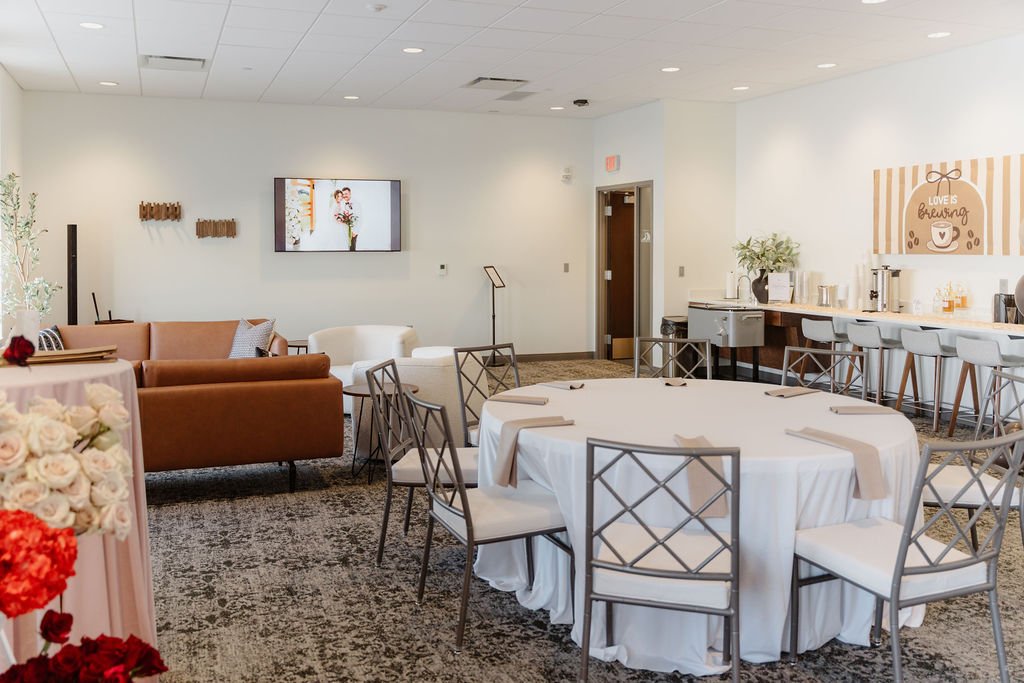 A decorated event space with round tables covered in white tablecloths, surrounded by metal chairs with white cushions, a sitting area with a brown sofa and white armchairs, a drink station with a counter and barstools, and floral arrangements, suita