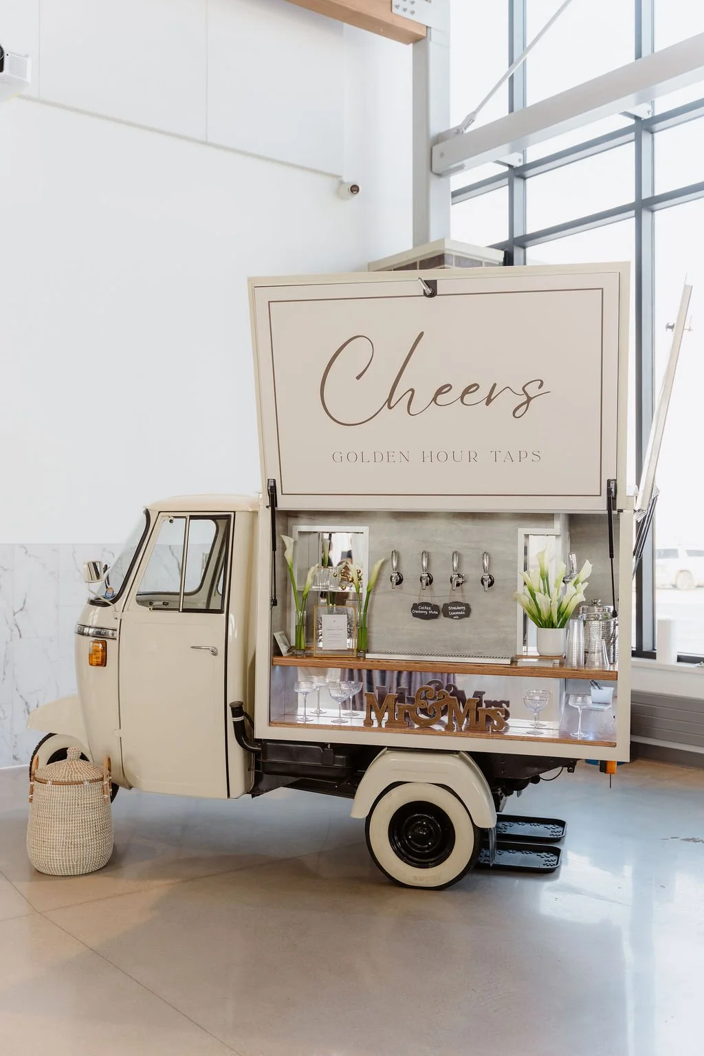 A mini vintage-style vehicle serving as a mobile bar with a sign that reads 'Cheers' and 'Golden Hour Taps' at an indoor event space.