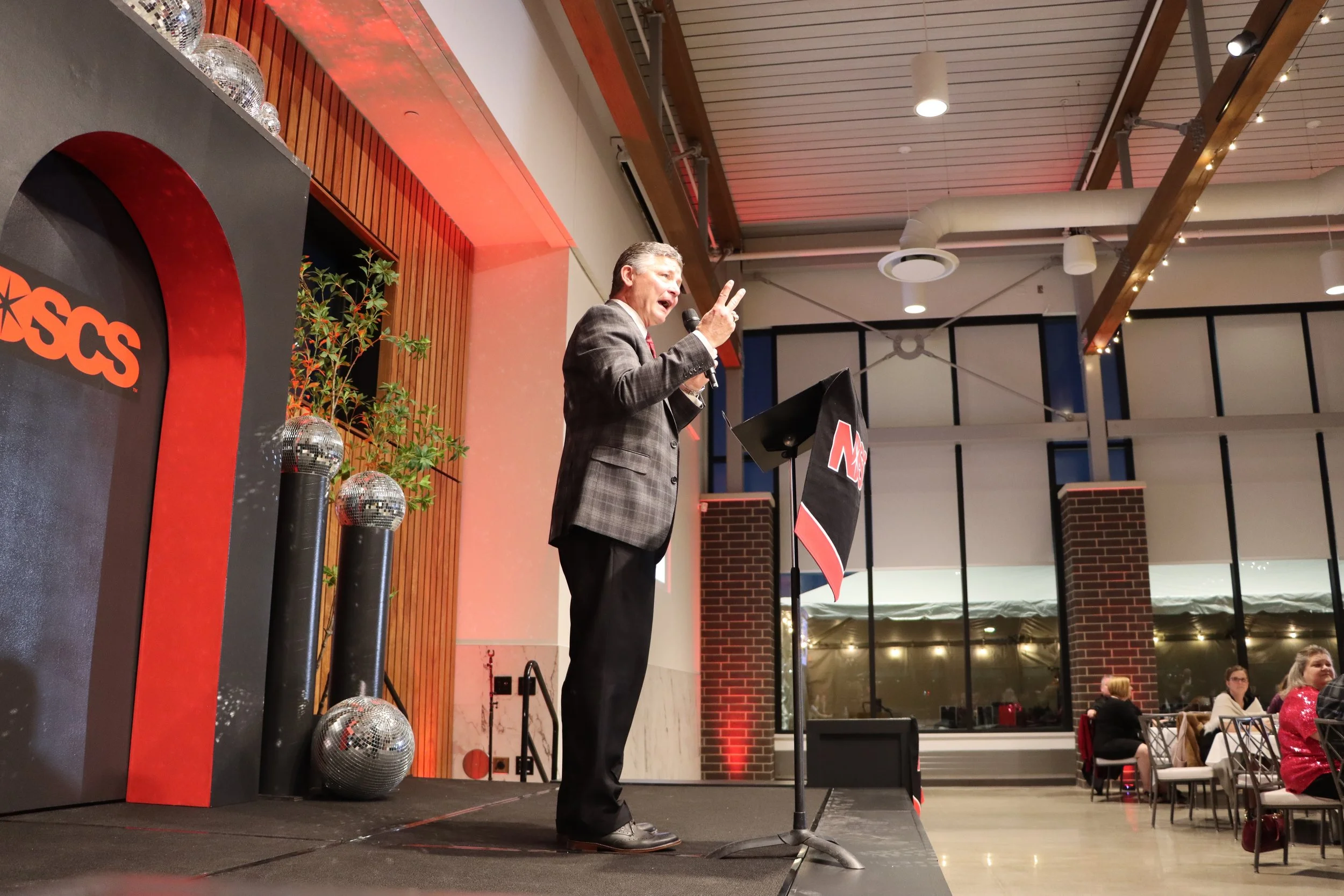 A man in a plaid suit giving a speech or presentation at an event in an indoor setting, with a microphone in his right hand and making a peace sign with his left hand. Multiple disco balls and a large sign with the partial word "SCS" are visible on t