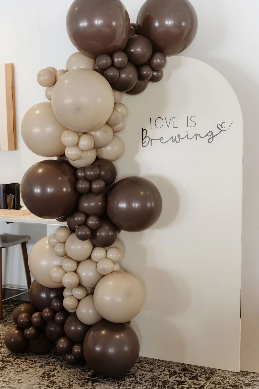 Balloon display with beige and brown balloons arranged in a column next to a white wall with the sign 'Love is Brewing' in black cursive and print text.