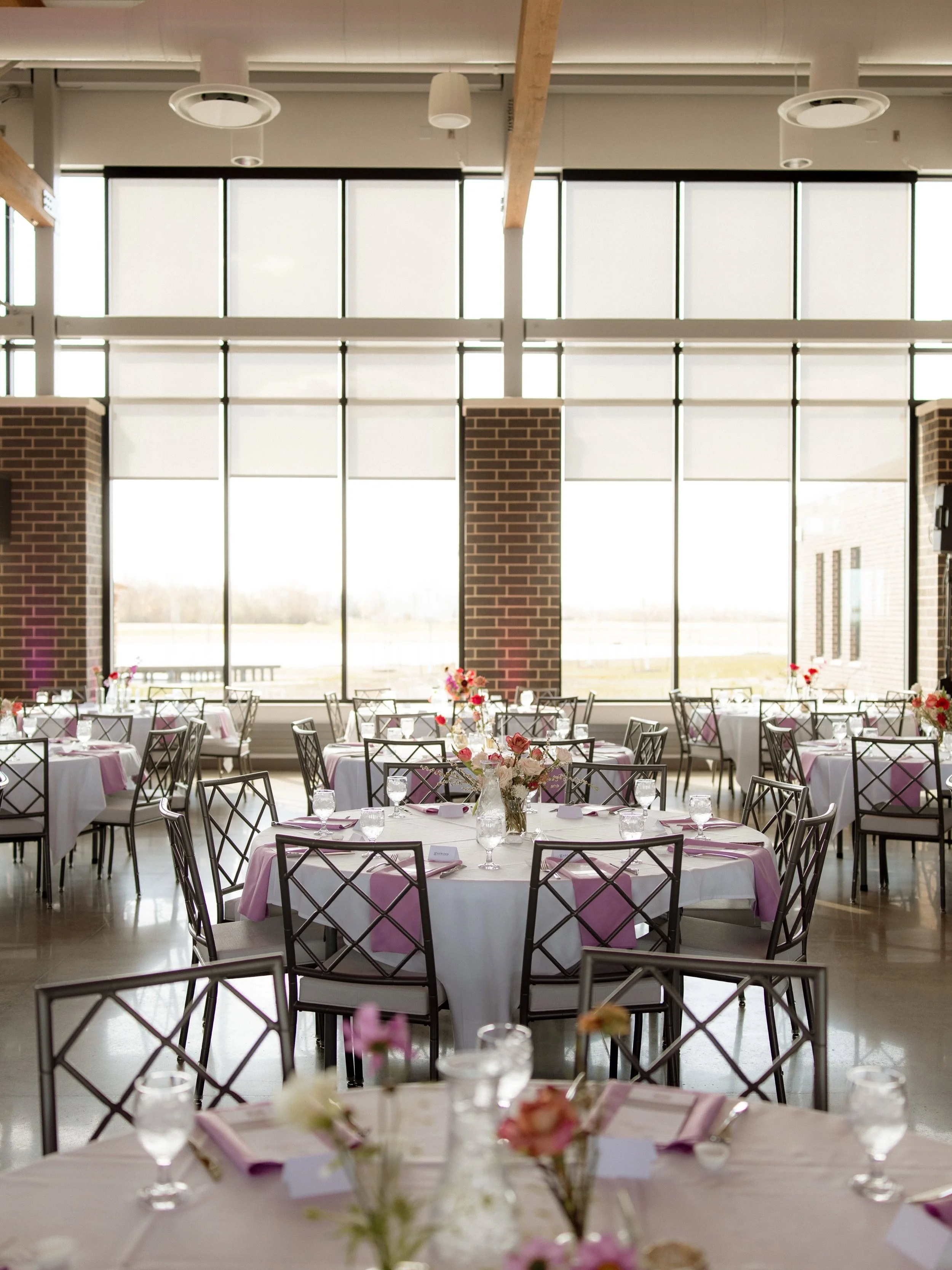 Event space with round tables covered in white and pink tablecloths, decorated with floral centerpieces, glassware, and place cards. Large windows with frosted glass panels in the background allow natural light into the room.