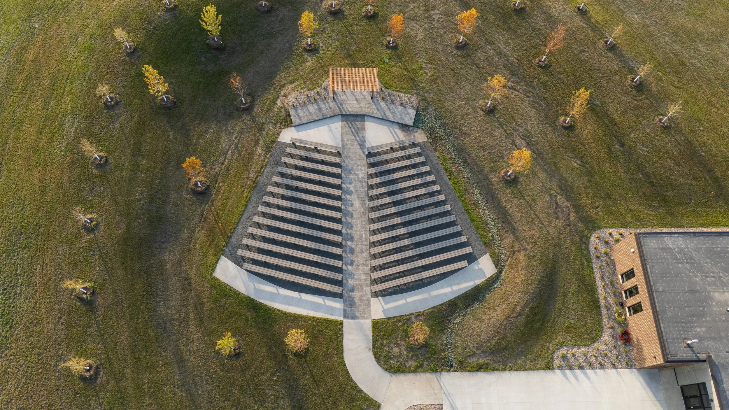An aerial view of a modern architectural structure with a curved concrete walkway leading to it, surrounded by evenly spaced young trees on a grassy area.