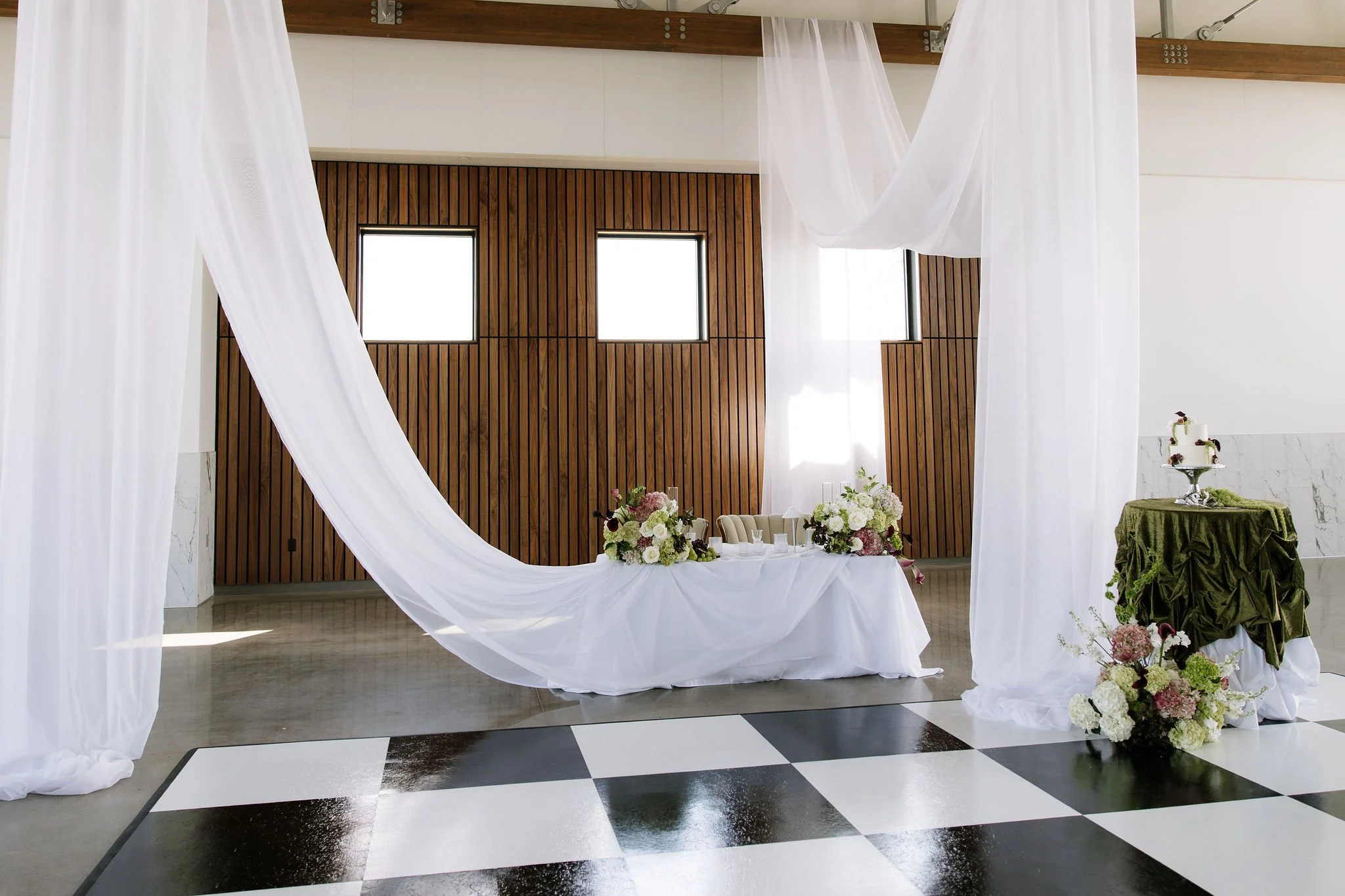 Wedding reception setup with a table and floral arrangements, draped white fabric, and a green-covered display with a Tiered cake, in a modern indoor venue with wood-paneled walls.