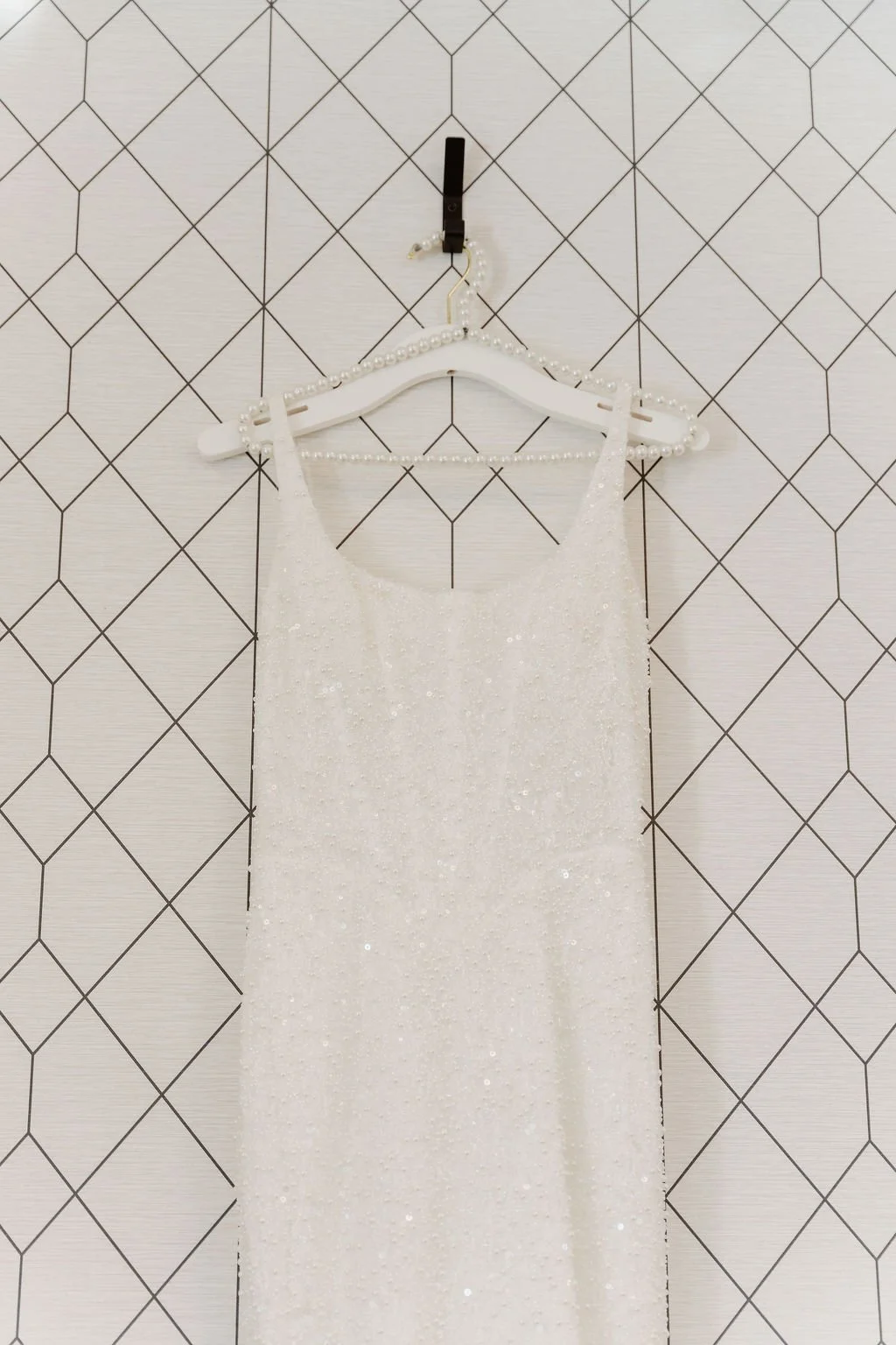 A white dress with sequins hangs on a pearl-white hanger attached to a black hook on a geometric-patterned wall.