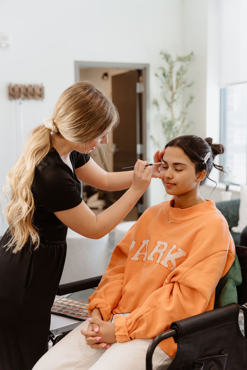 Makeup artist applying makeup to a young woman sitting in a wheelchair in a brightly lit room.