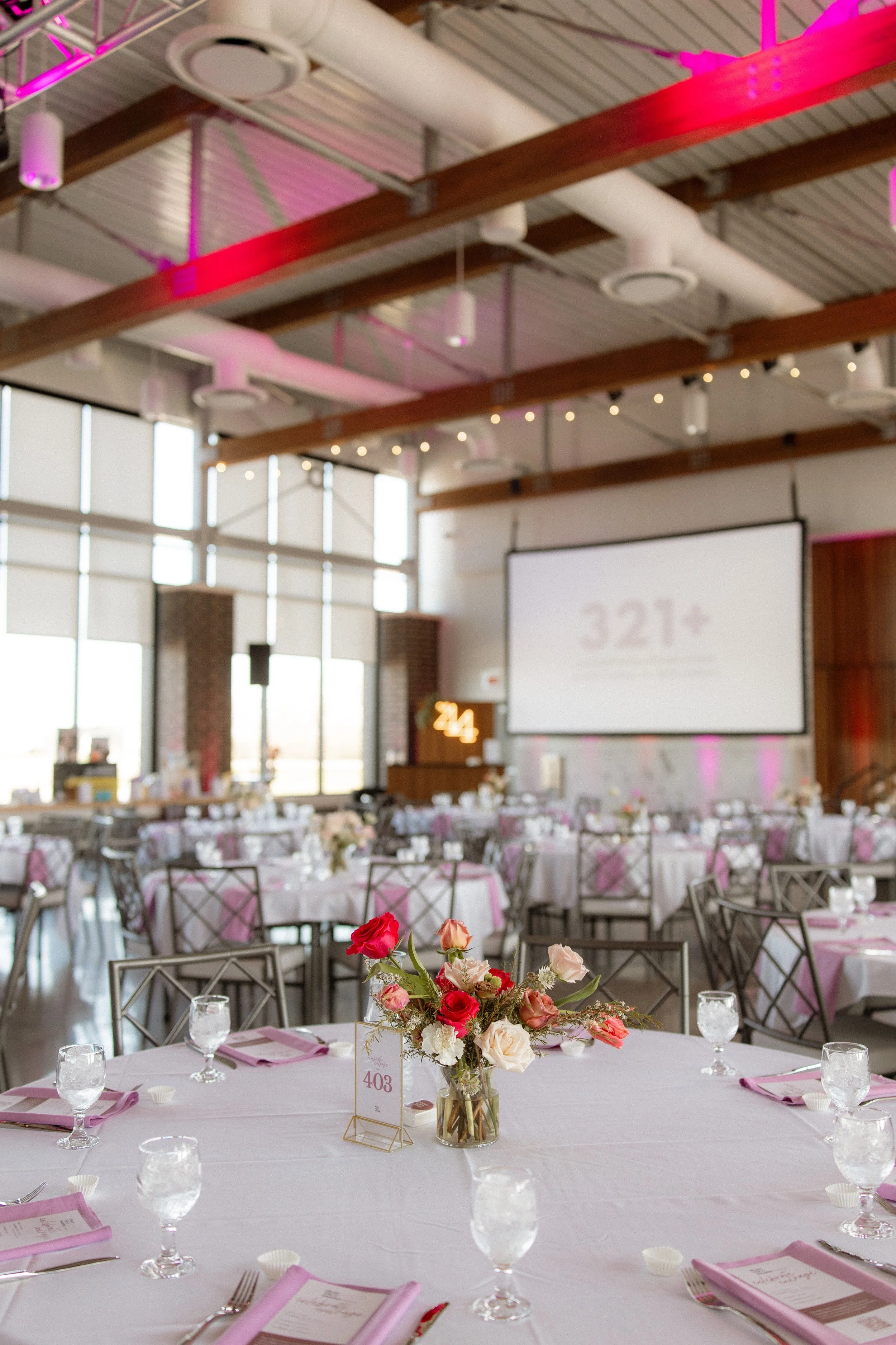 A decorated banquet hall with round tables covered in white tablecloths, pink napkins, and floral centerpieces with pink, red, and white flowers, set for a wedding or event.