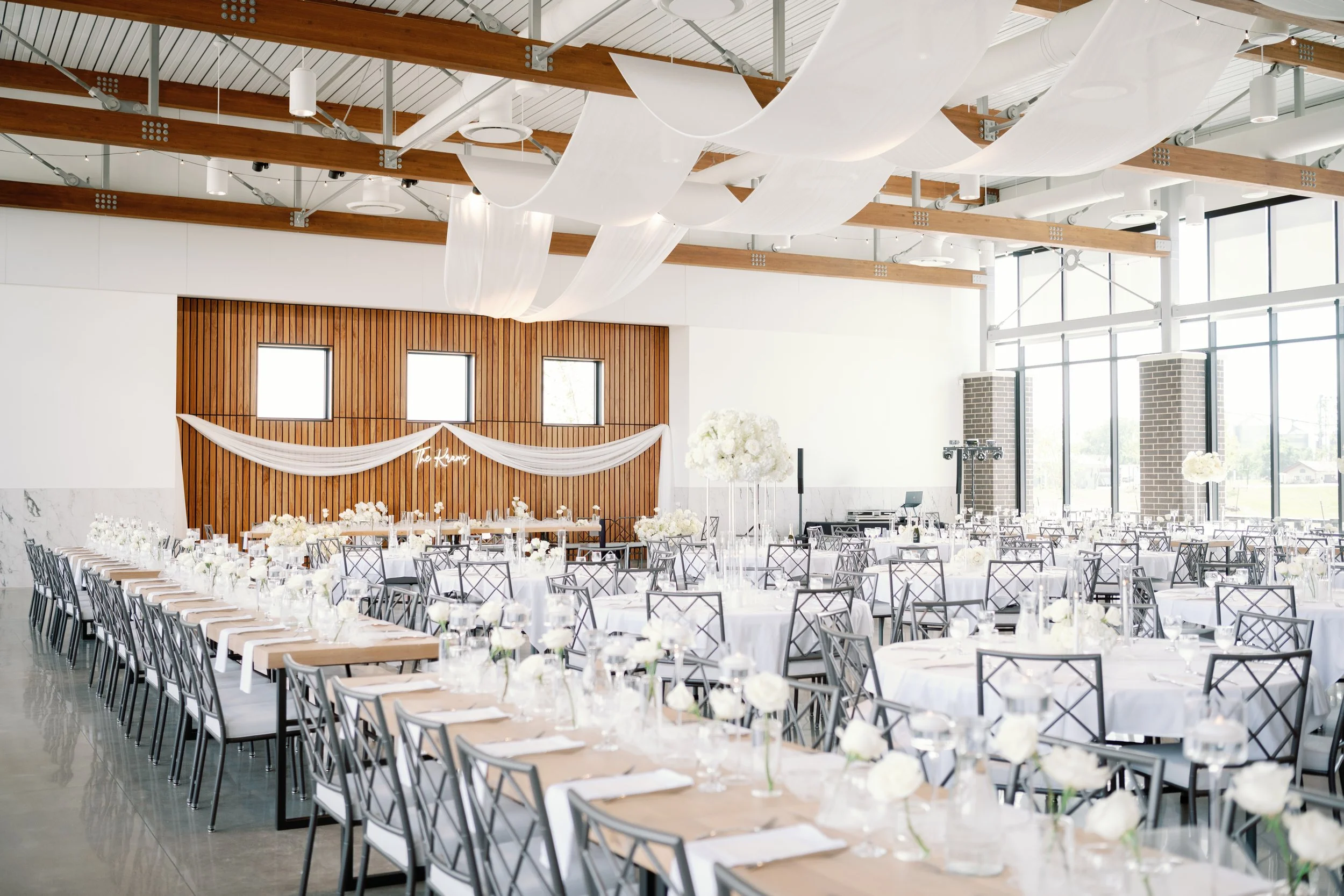 A large, bright event hall with multiple round and rectangular tables covered with white tablecloths and decorated with white flower arrangements. The hall features high ceilings with exposed beams and large windows along one wall, allowing natural l
