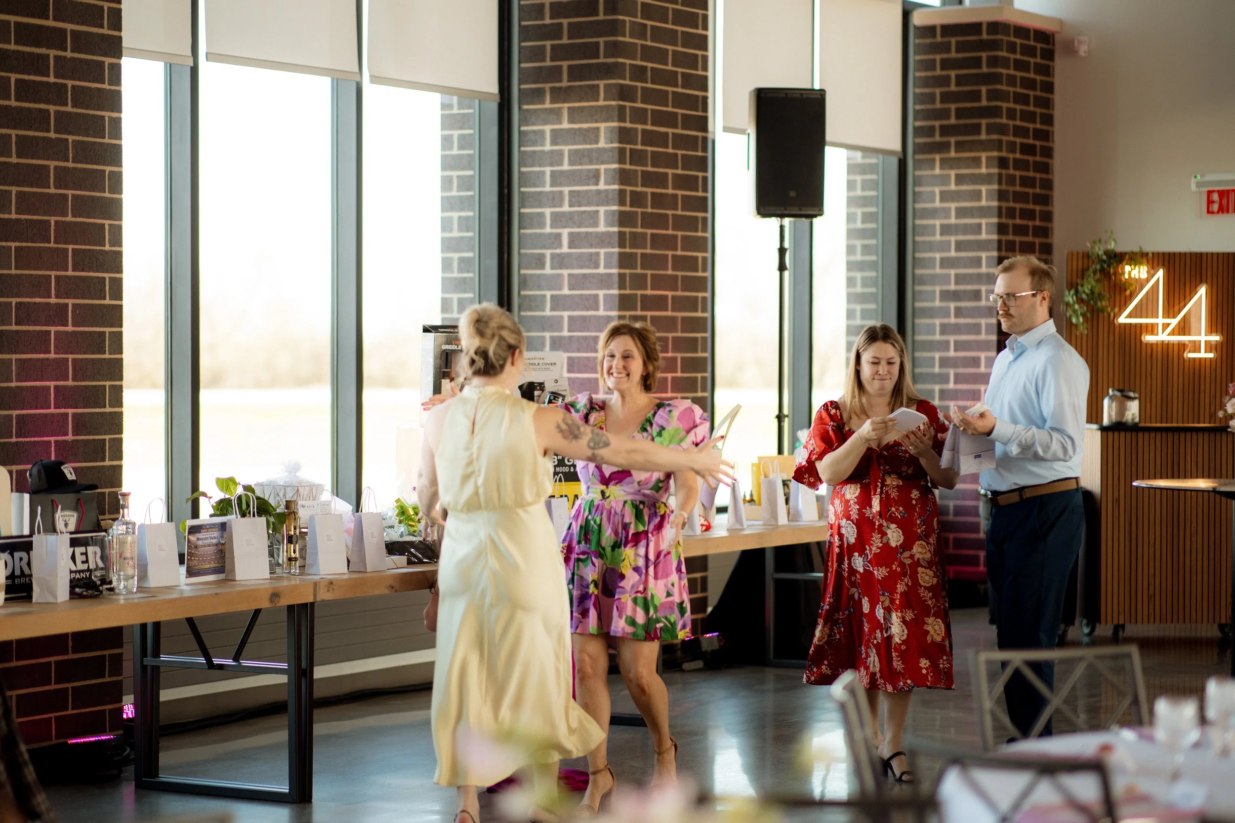 People standing at a table with gift bags in a room with large windows and brick walls. A woman in a yellow dress interacts with women in floral and red dresses. A man in a light blue shirt holds papers nearby.