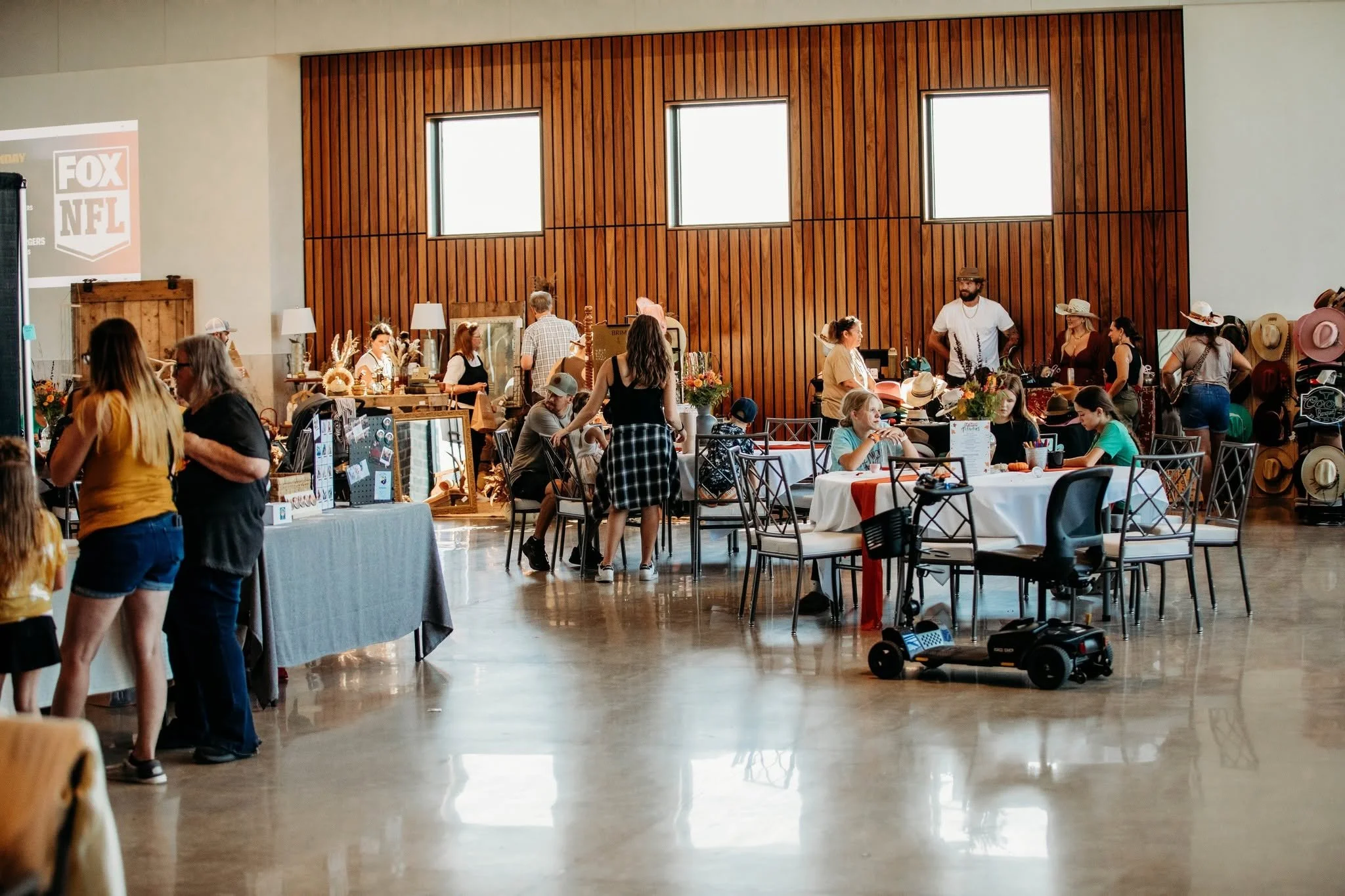 Indoor event space with people browsing vendor booths, some sitting at tables, wooden wall with large windows, and a vending machine.
