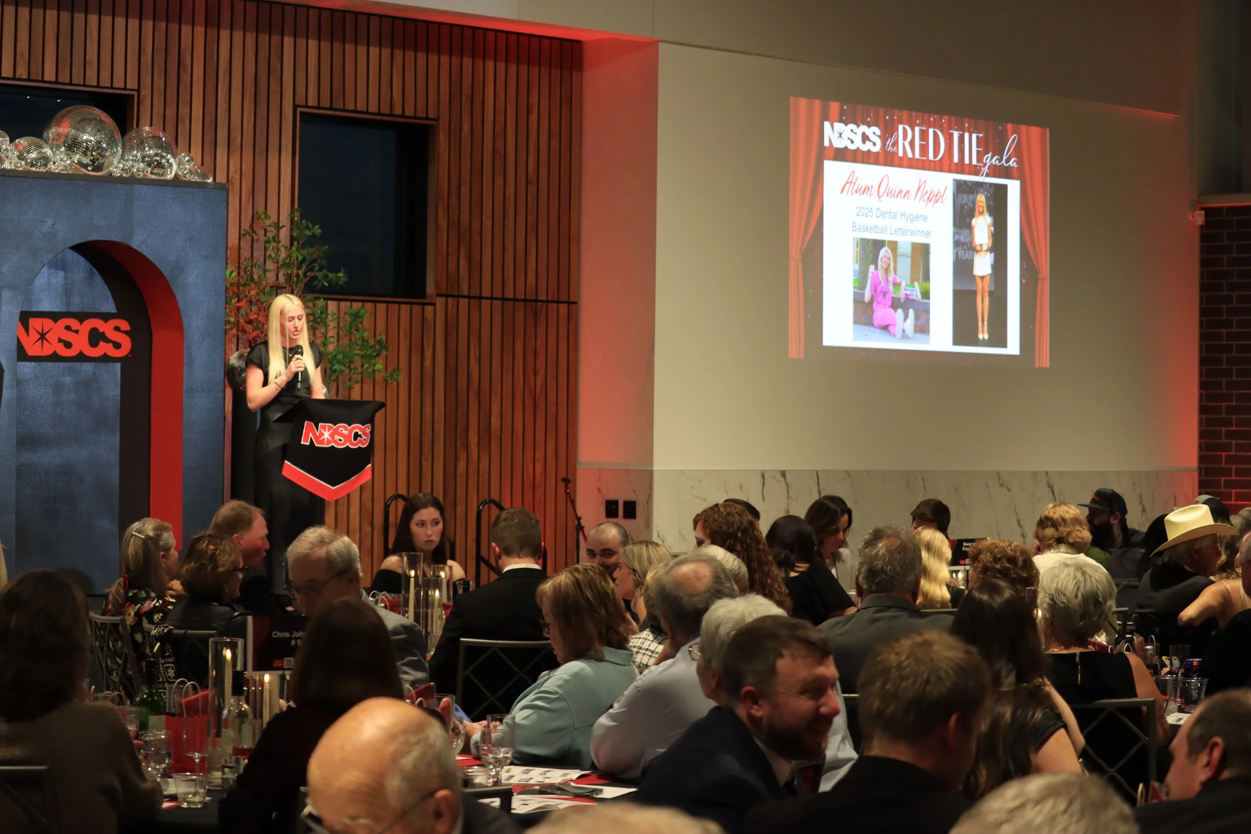 A woman with long blonde hair is speaking at a podium with the NSCS logo during a gala event. There is a large projection screen displaying a slide about Alum Quinn Neppi, a 2025 dental hygiene graduate and basketball letterwinner. The audience consi