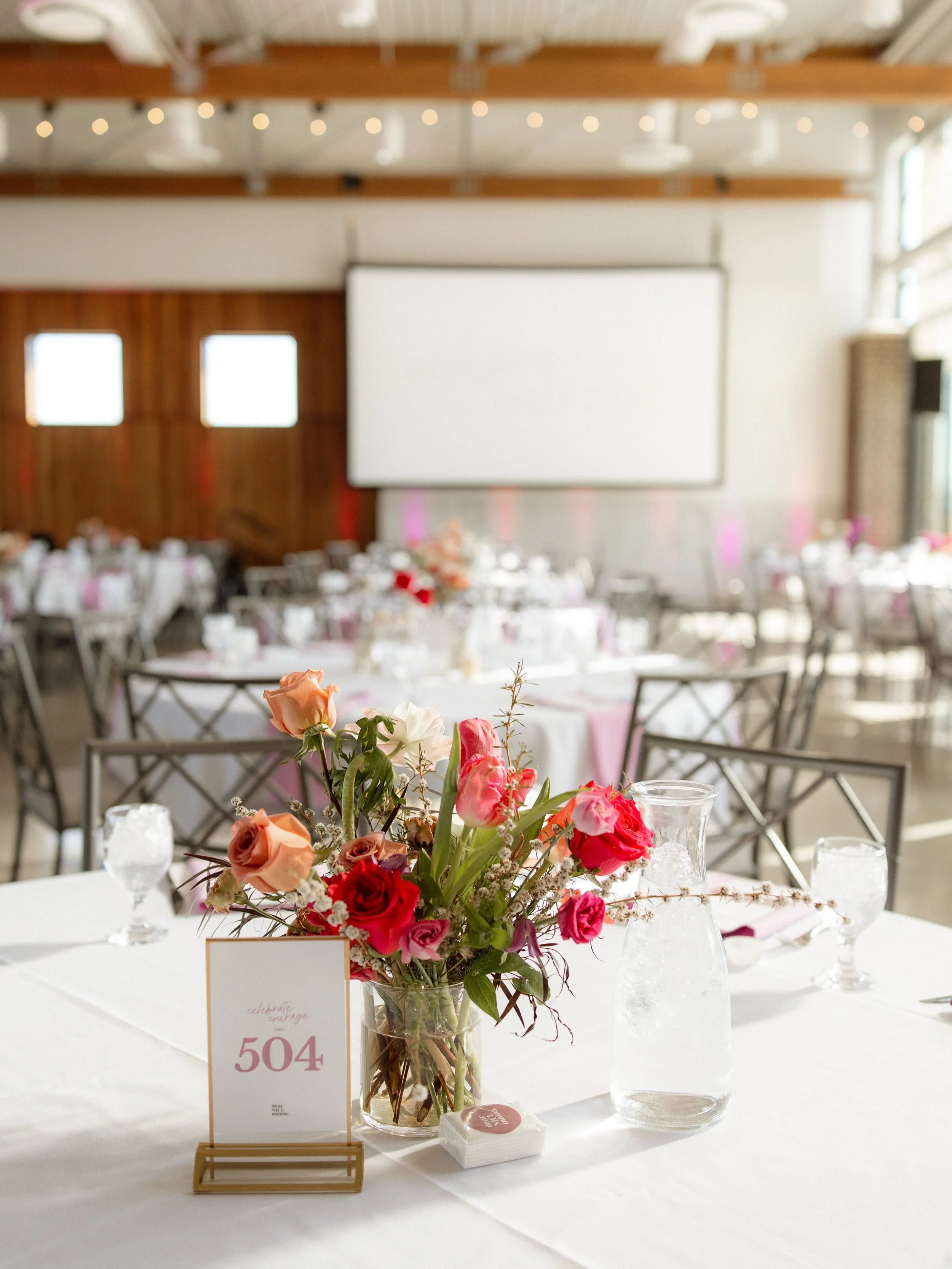 A table centerpiece with a bouquet of pink, peach, and red roses in a glass vase, surrounded by glass votive candle holders, on a white tablecloth at an event venue.