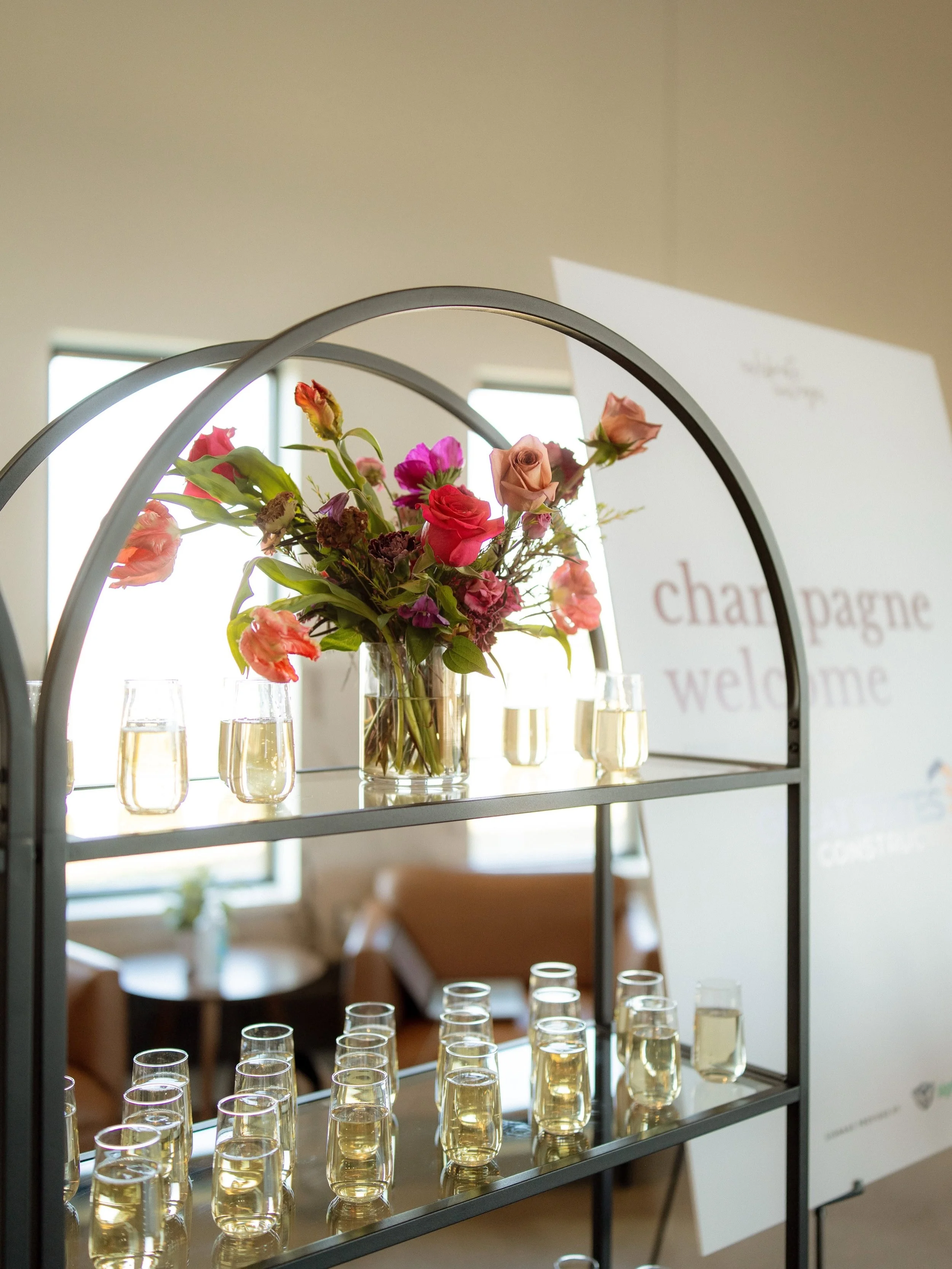 A metal display stand with three shelves holding a bouquet of pink and peach roses and other flowers in a clear glass vase on the top shelf, surrounded by clear glasses filled with champagne or white wine, in a bright room with windows and a white wa