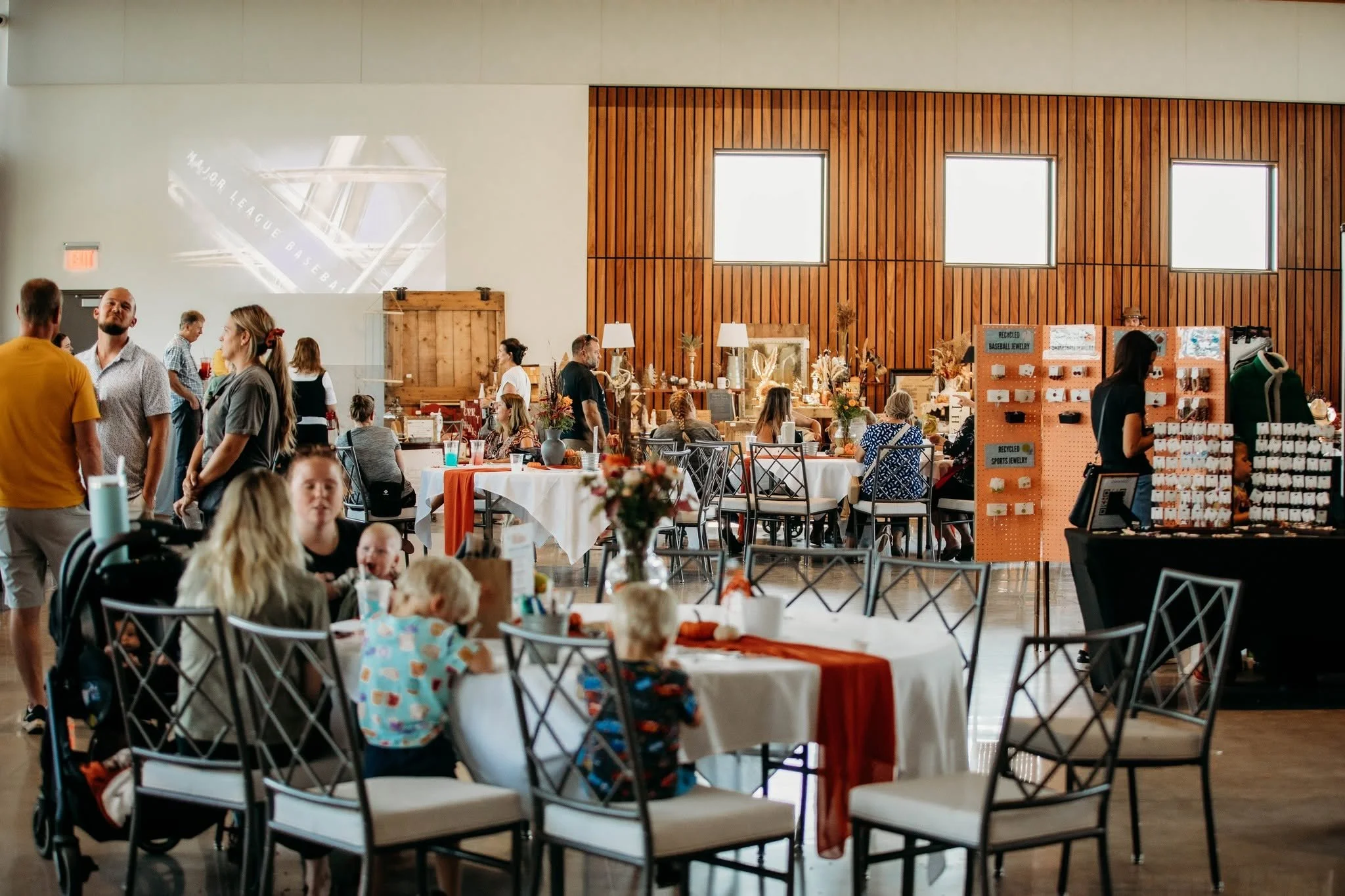 People dining and socializing in a decorated indoor event space with tables, chairs, and a wooden wall with windows.