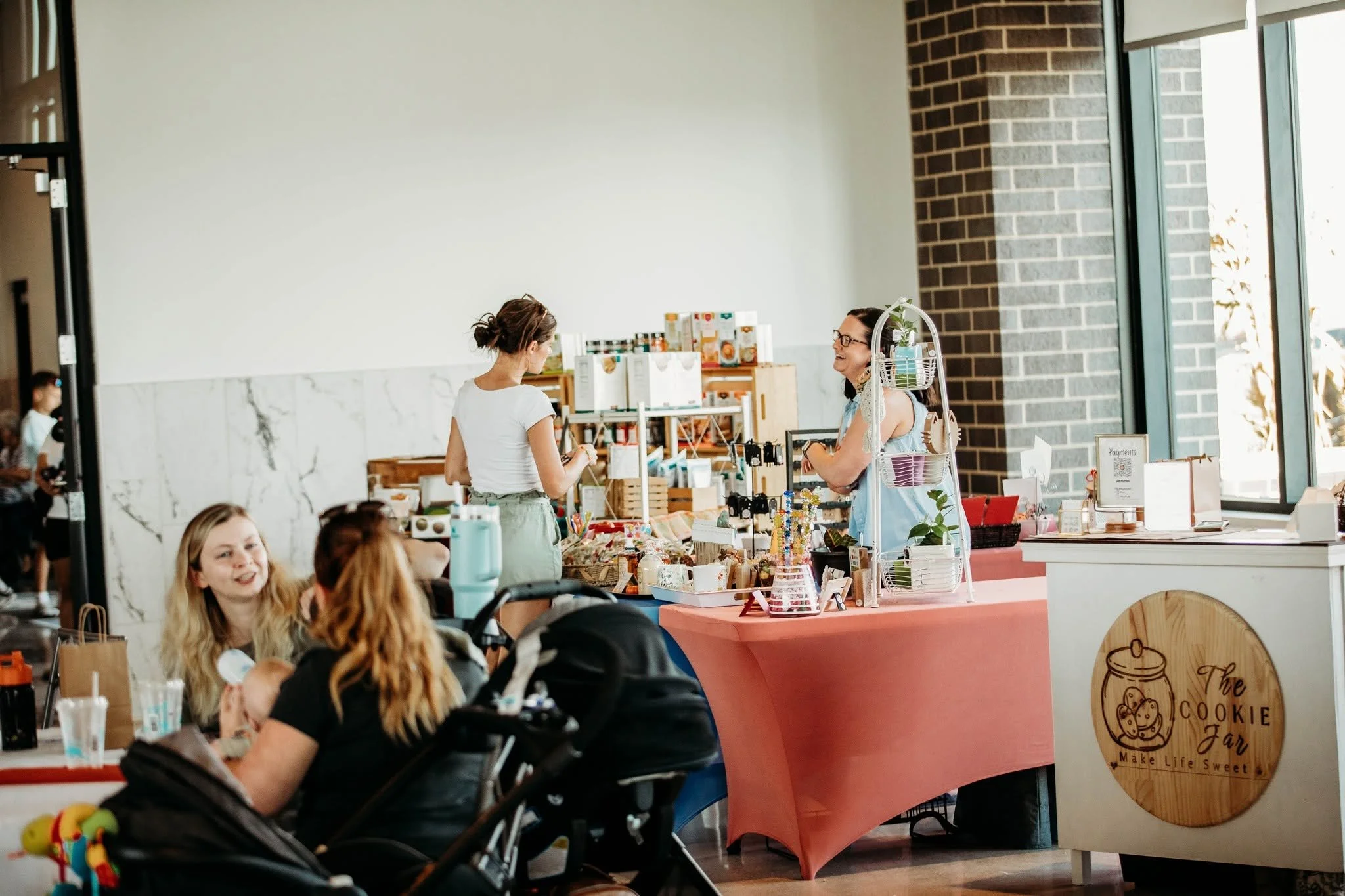 Two women stand behind a table at a vendor booth in a shopping mall, exchanging conversation, with corner of a sign that reads "The Cookie Jar" on the front of the booth, and shelves with various items in the background.