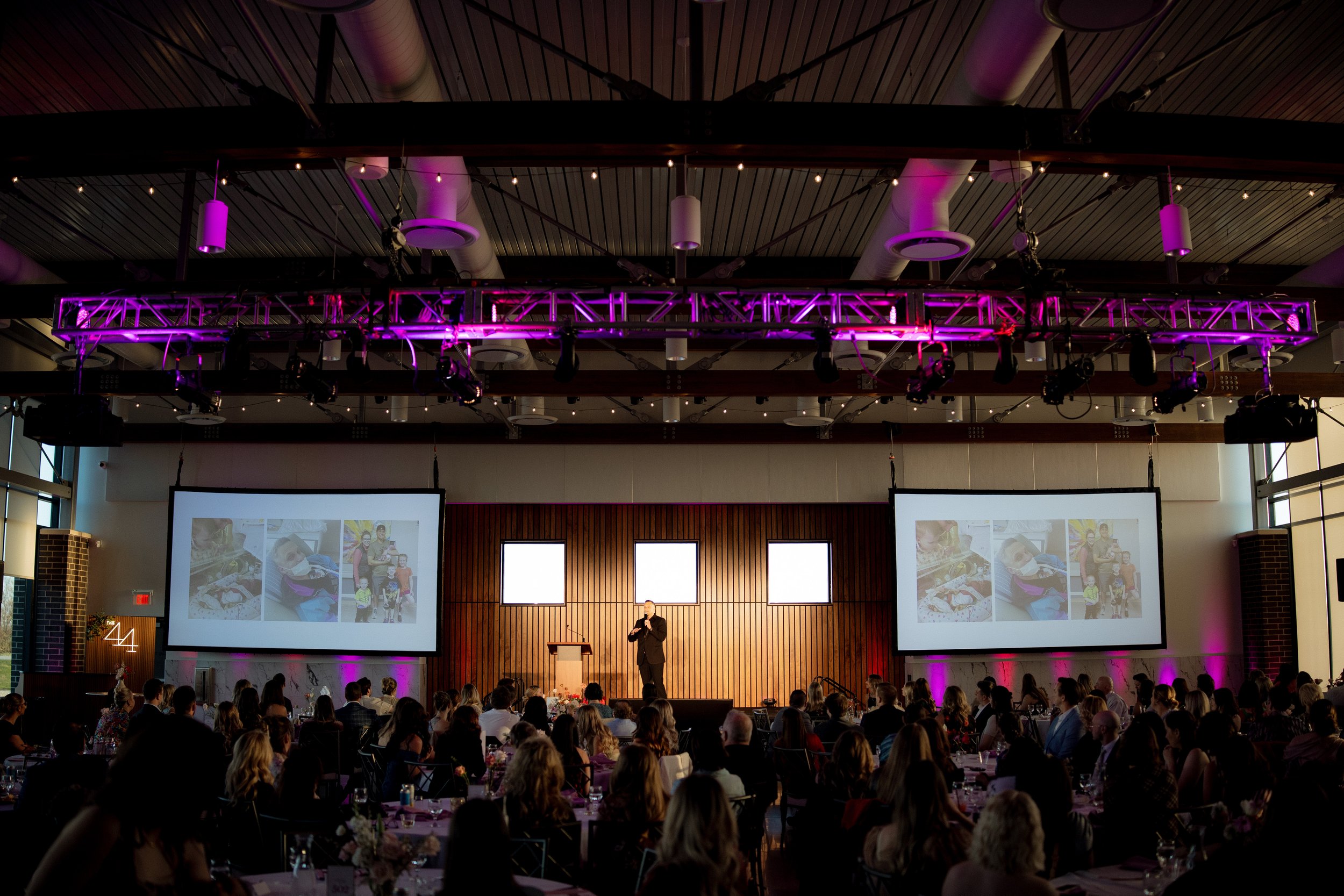Large indoor event space with a stage, two presentation screens showing photos of children, and a speaker standing on the stage. The audience sits at round tables, and there are purple and pink stage lights illuminating the ceiling.