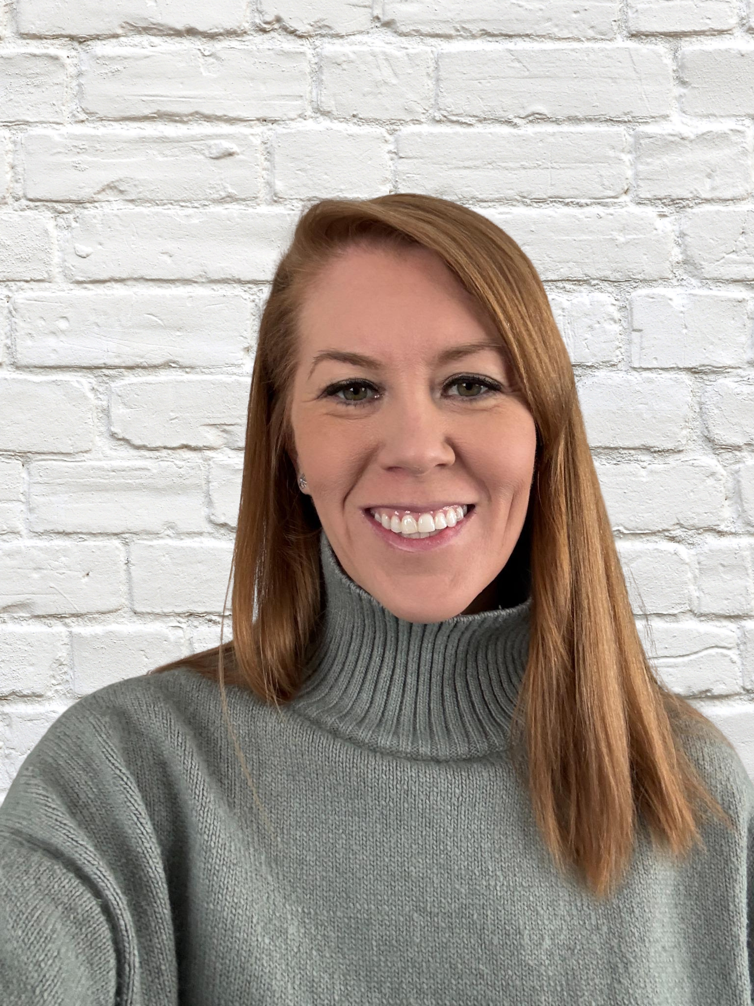 Person smiling in front of white brick wall, wearing a gray turtleneck sweater.
