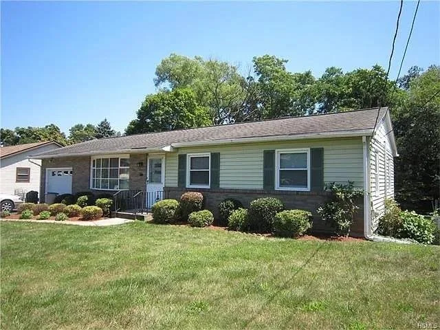 Small single-story house with light green siding, white trim, and a shingled roof. The front yard features neatly trimmed bushes and a grassy lawn. A concrete path leads to the front door. Trees and a garage are in the background.