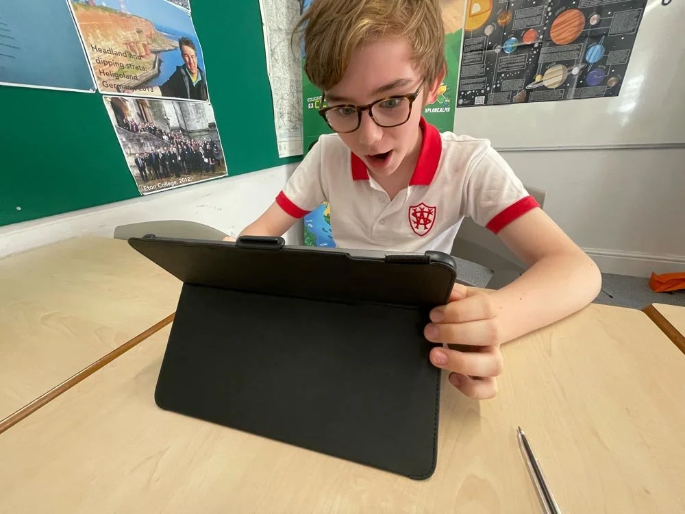 A young boy with glasses sitting at a classroom desk, looking at a tablet with an expression of surprise or excitement. He is wearing a white polo shirt with red accents and a crest on the chest.