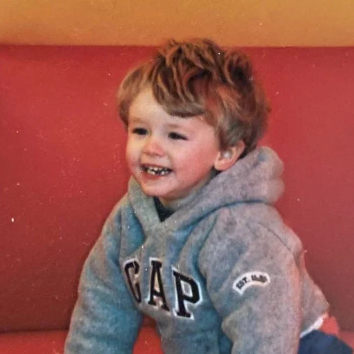 A young boy with curly red hair smiling and wearing a gray GAP hoodie, sitting in front of a pink wall.