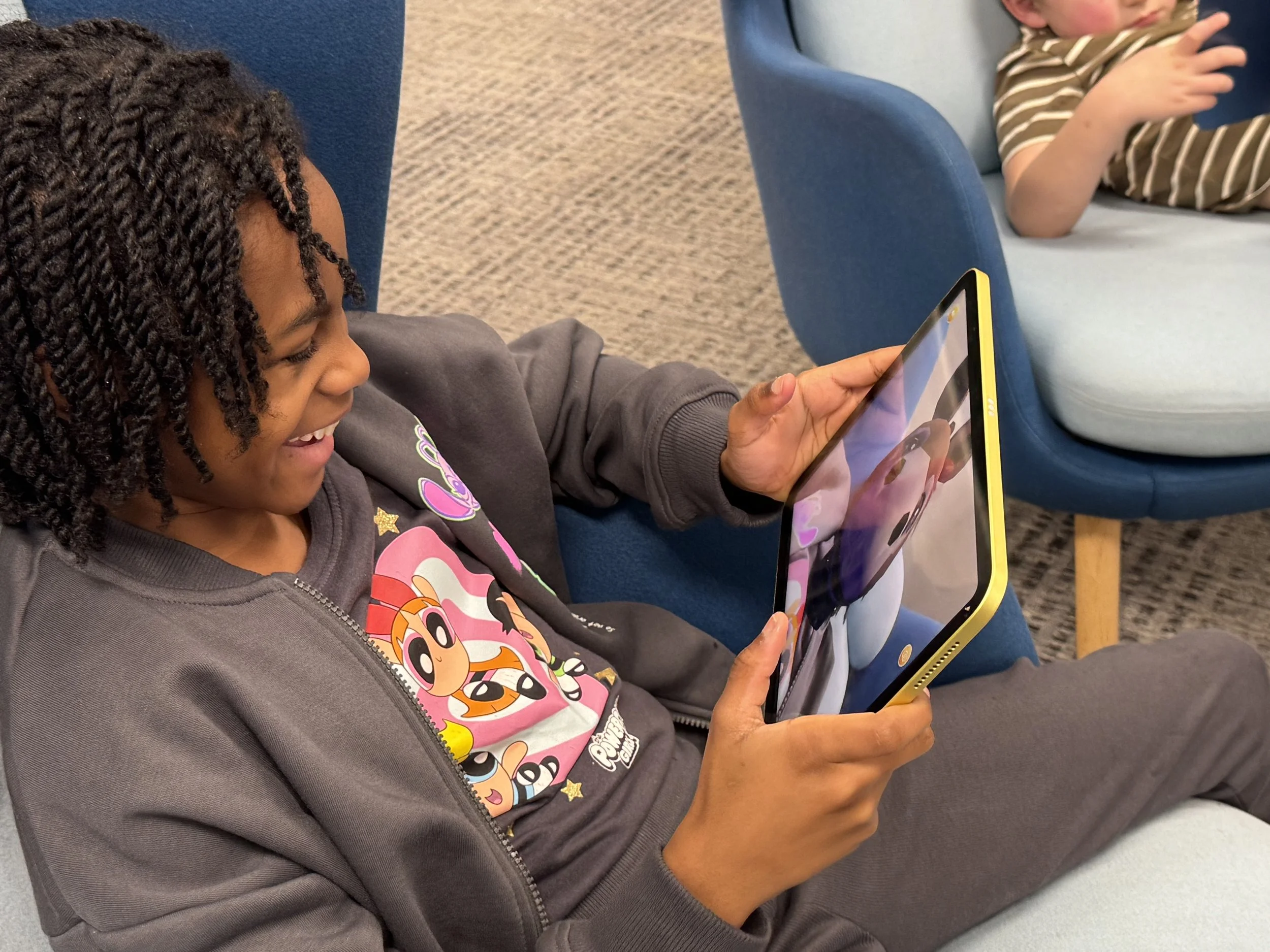 A young girl with curly hair smiling while playing a game on a yellow tablet, sitting on a blue chair with another child nearby.
