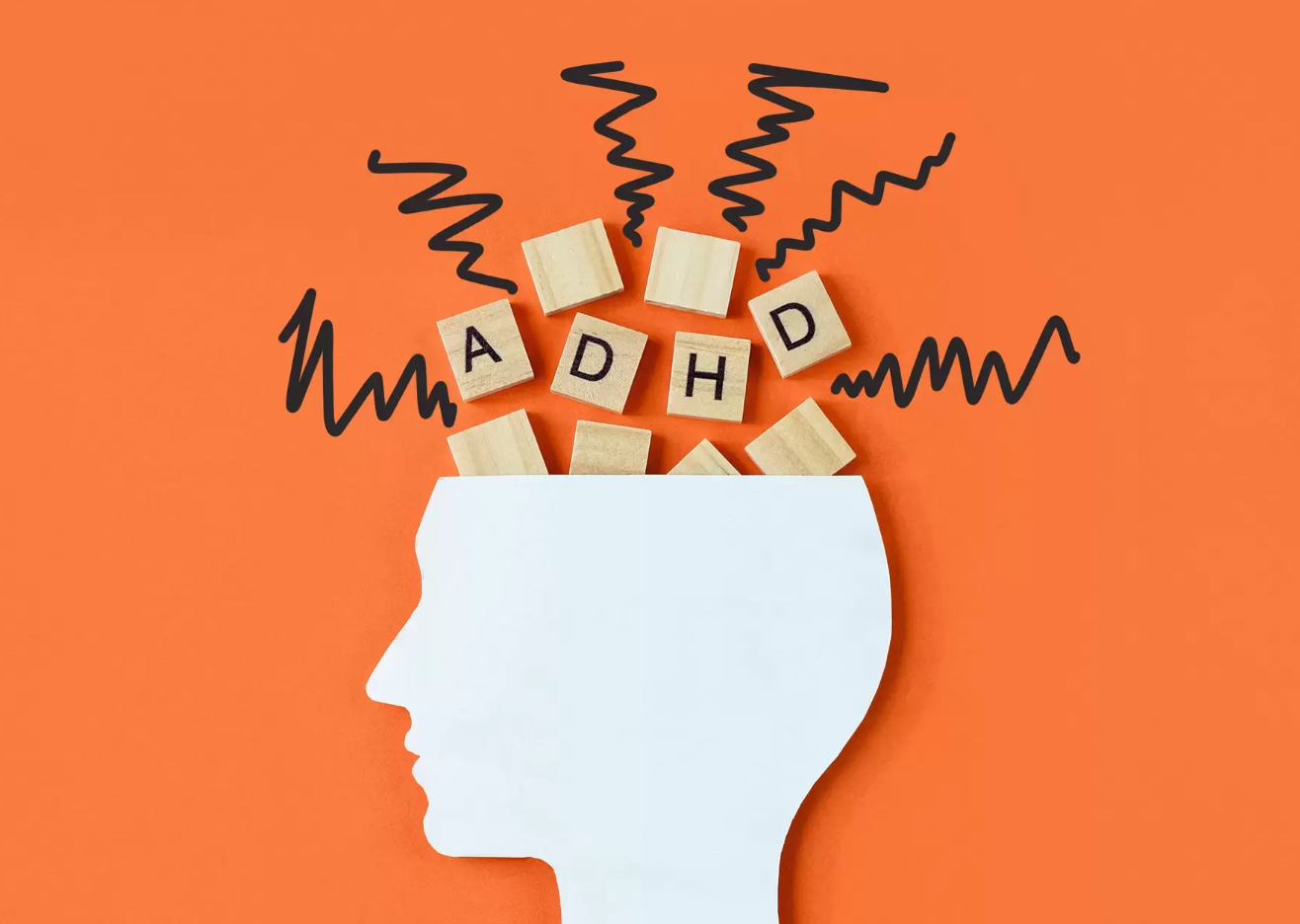 Orange background. Side silhouette of a person face with wooden letters spelling out ADHD and squiggly lines coming out the top of the head