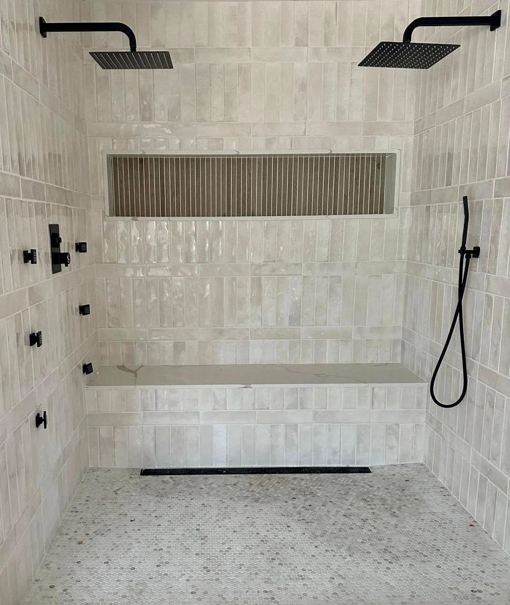 A modern shower area with beige tiles, two black rainfall showerheads, black fixtures, a black hand-held shower wand, a built-in tiled bench, a long horizontal niche with black grill, and a small drain on the floor.