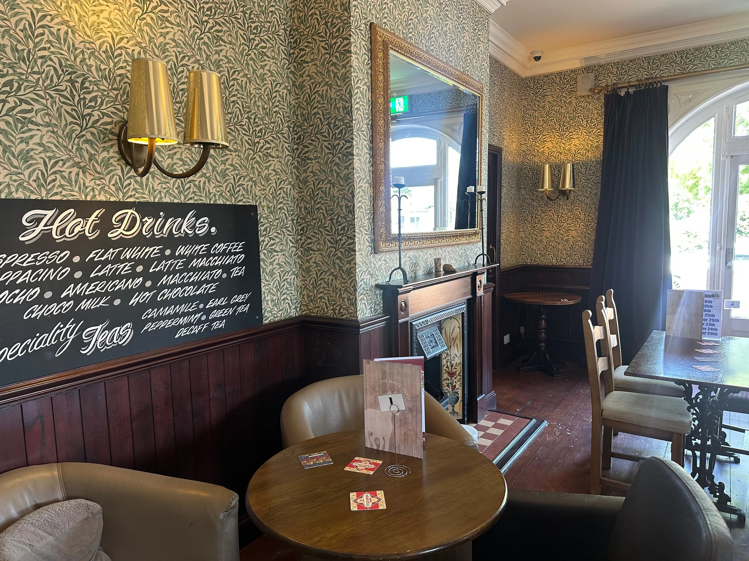 Interior of a cozy café with a blackboard menu listing hot drinks and specialty teas, vintage wallpaper, gold wall sconces, a large mirror, a fireplace with a decorative mantle, wooden furniture, an emergency exit sign, and sunlight coming through la