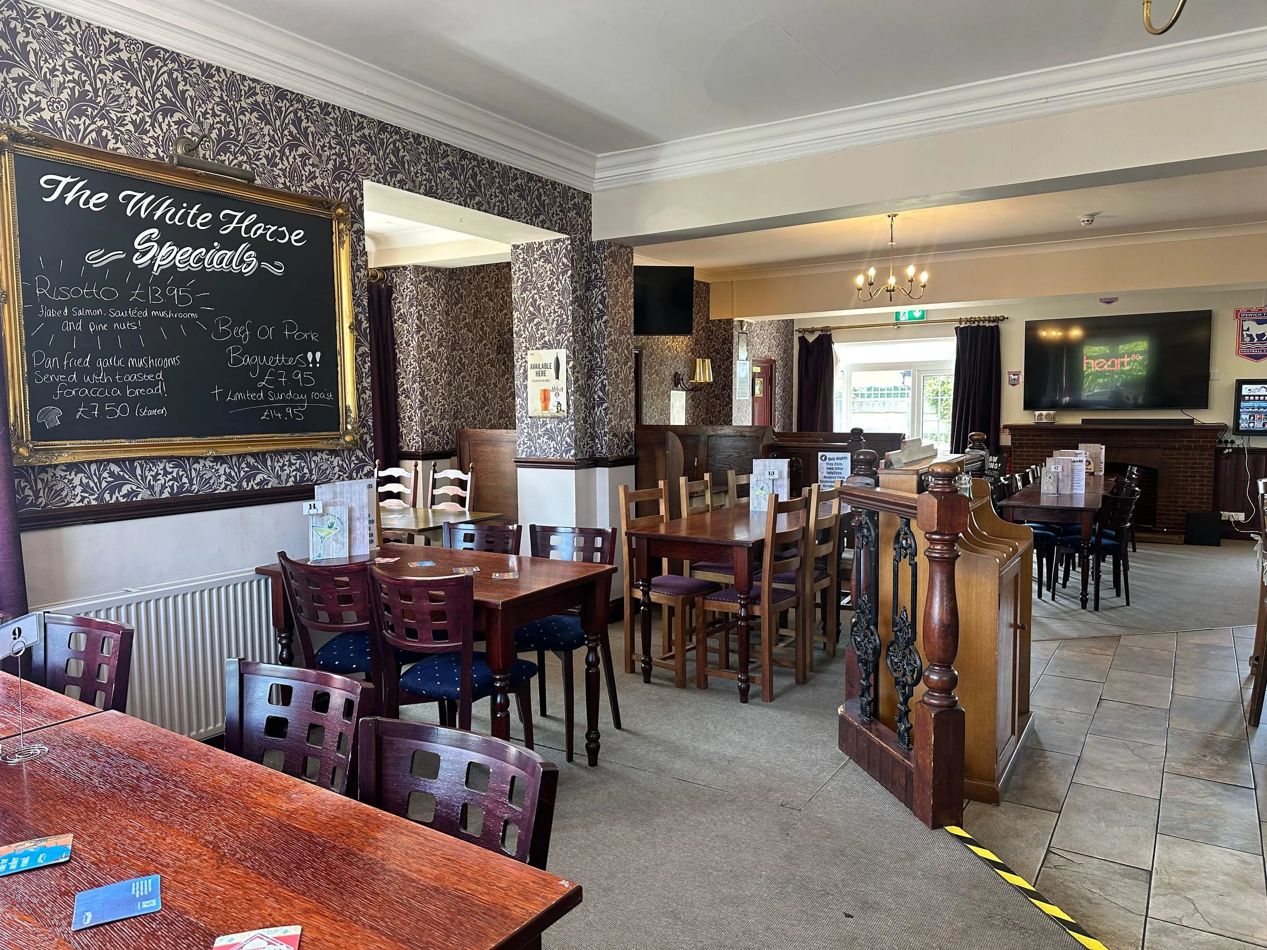Interior of a cozy restaurant with dark wood tables and chairs, patterned wallpaper, a large chalkboard menu listing specials, a flat-screen TV on the wall, and a view of the entrance with glass doors.