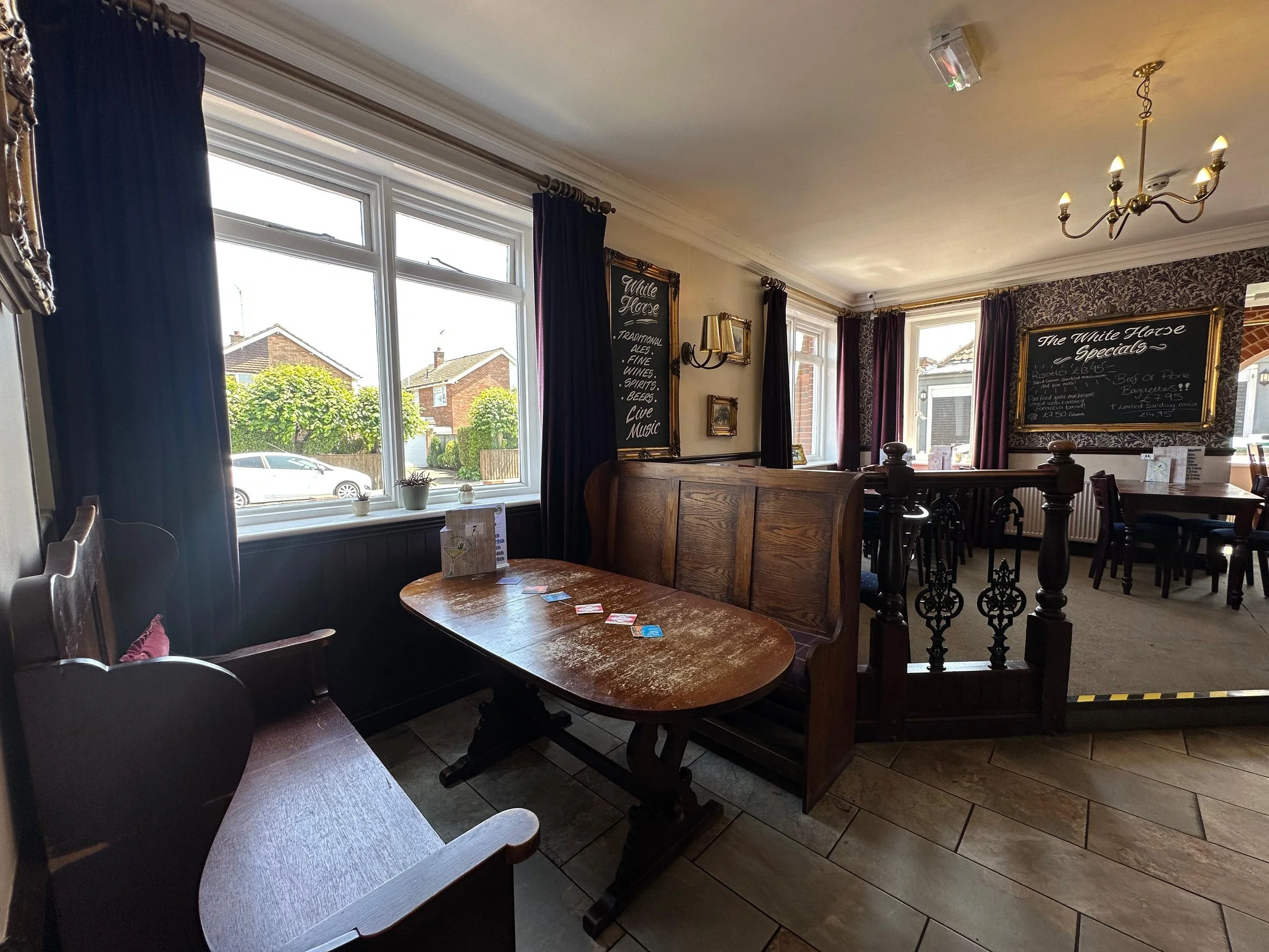Interior of a cozy pub or restaurant with large windows, dark curtains, wooden furniture, a chandelier, and a blackboard menu on the wall.