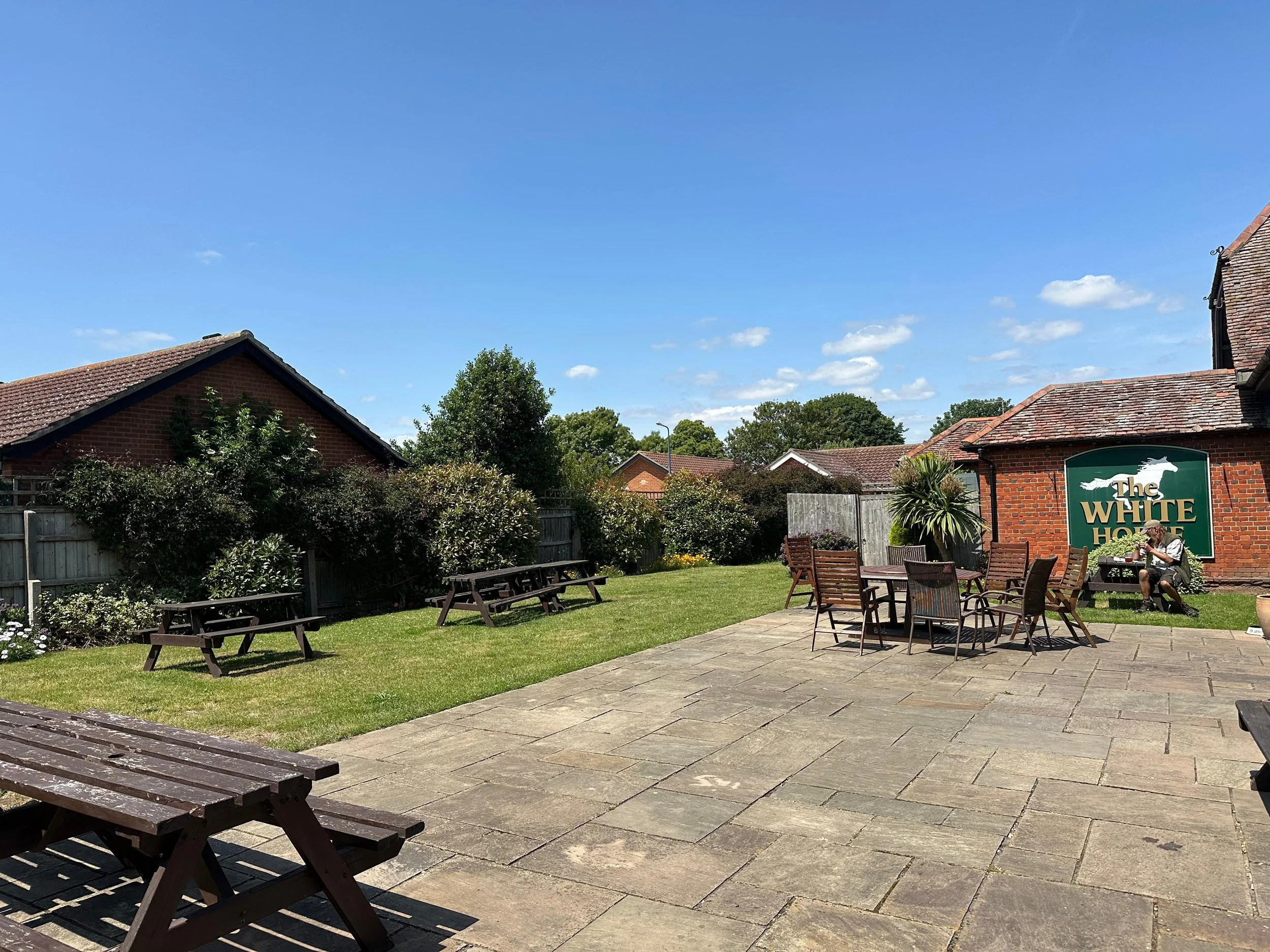 Patio area with wooden picnic tables and outdoor dining set, green lawn, bushes, trees, brick buildings, and a large sign for The White Horse under a blue sky with some clouds.