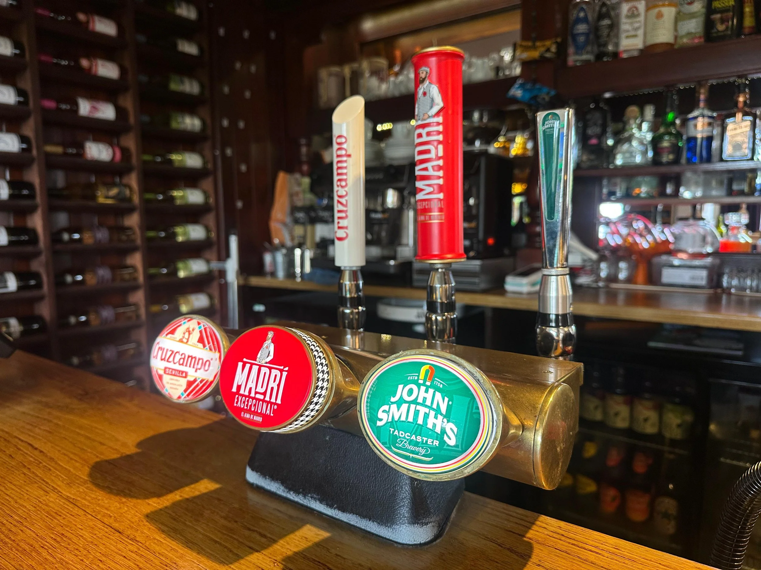 Beer tap handles at a bar with three different beers: Cruzcampo, Madri, and John Smith's, in a cozy, well-stocked bar setting.