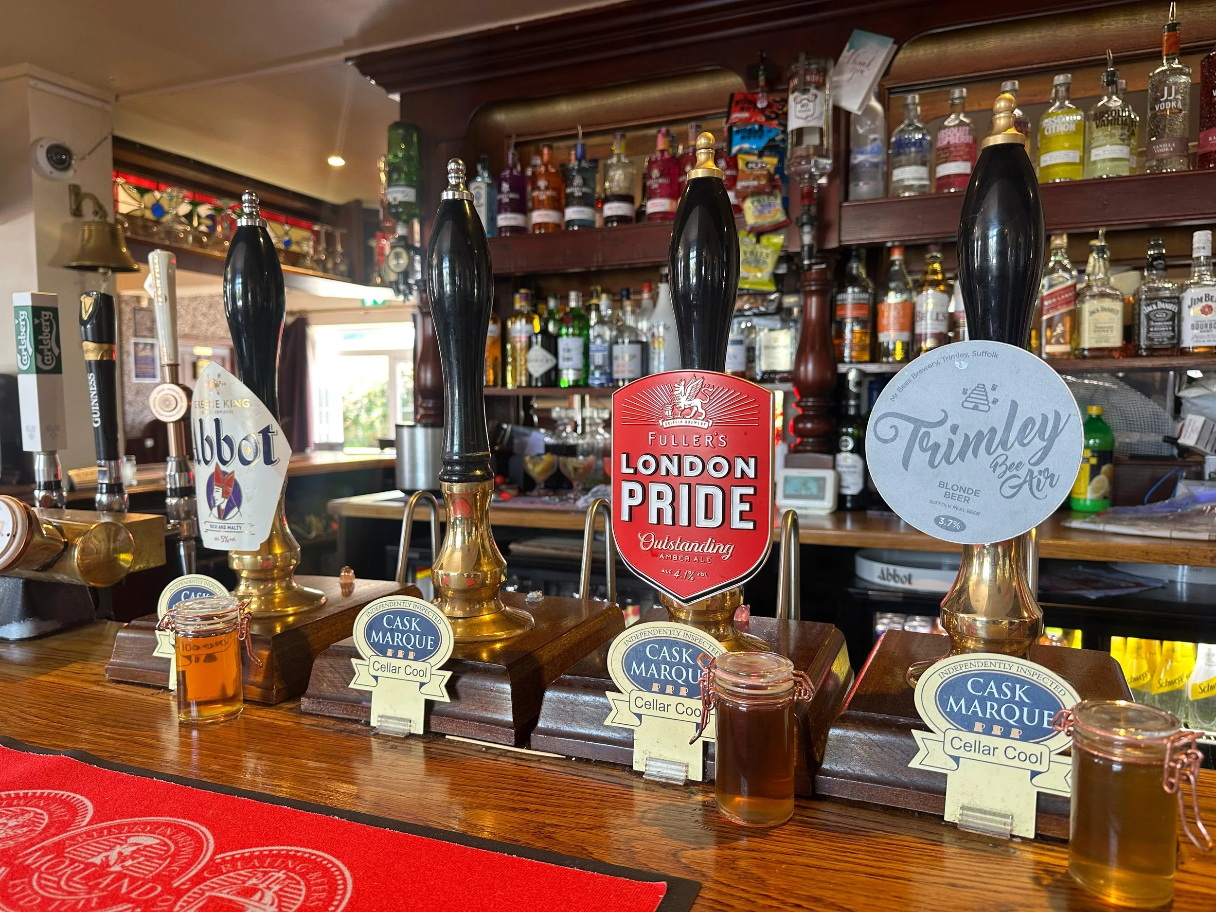 Bar counter with three beer taps labeled Abbot, Fuller’s London Pride, and Trimley Bee Air, with small tasting glasses filled with beer in front. Shelves stocked with various bottles of liquor are in the background.