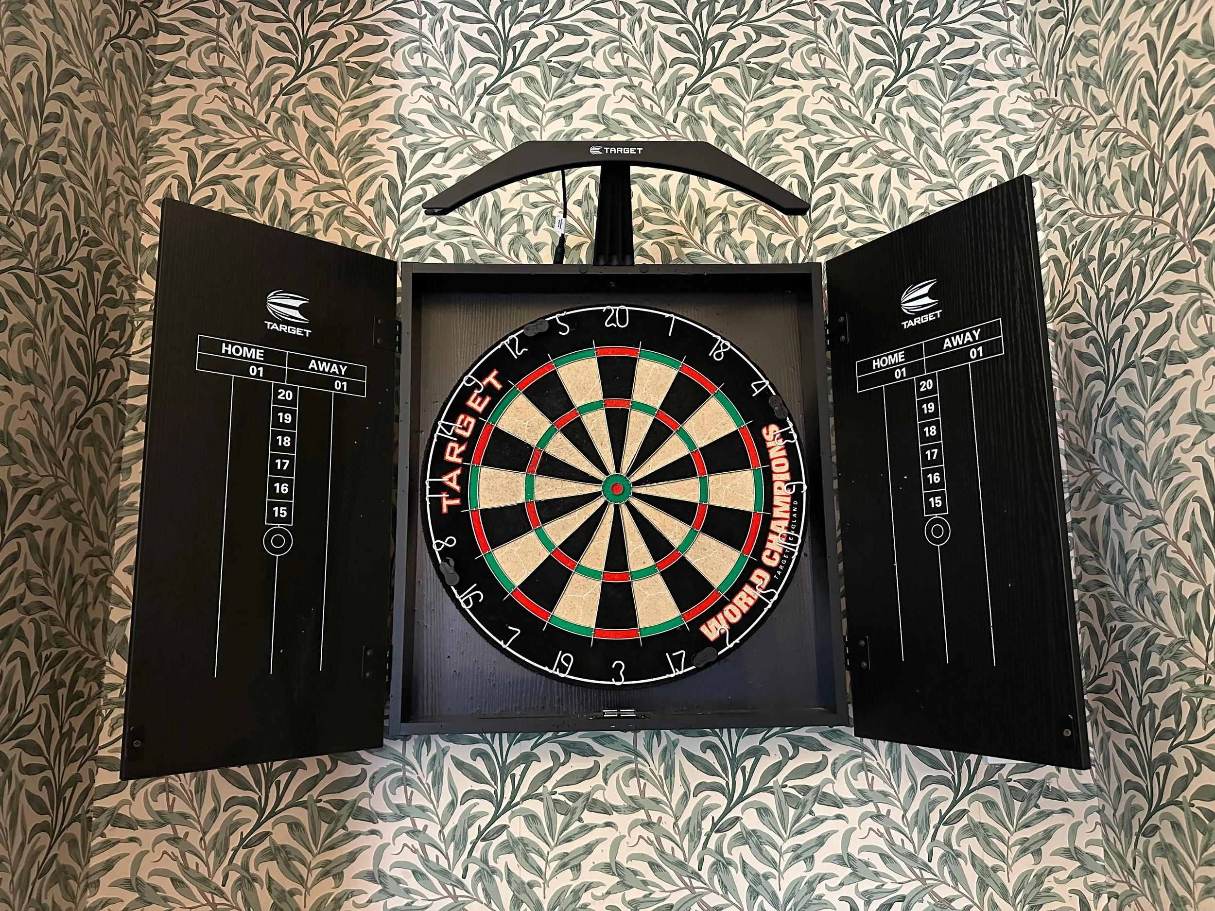 A mounted electronic dartboard with a circular target in the center, surrounded by a black cabinet with closed doors, on a wall with leafy green and beige wallpaper.