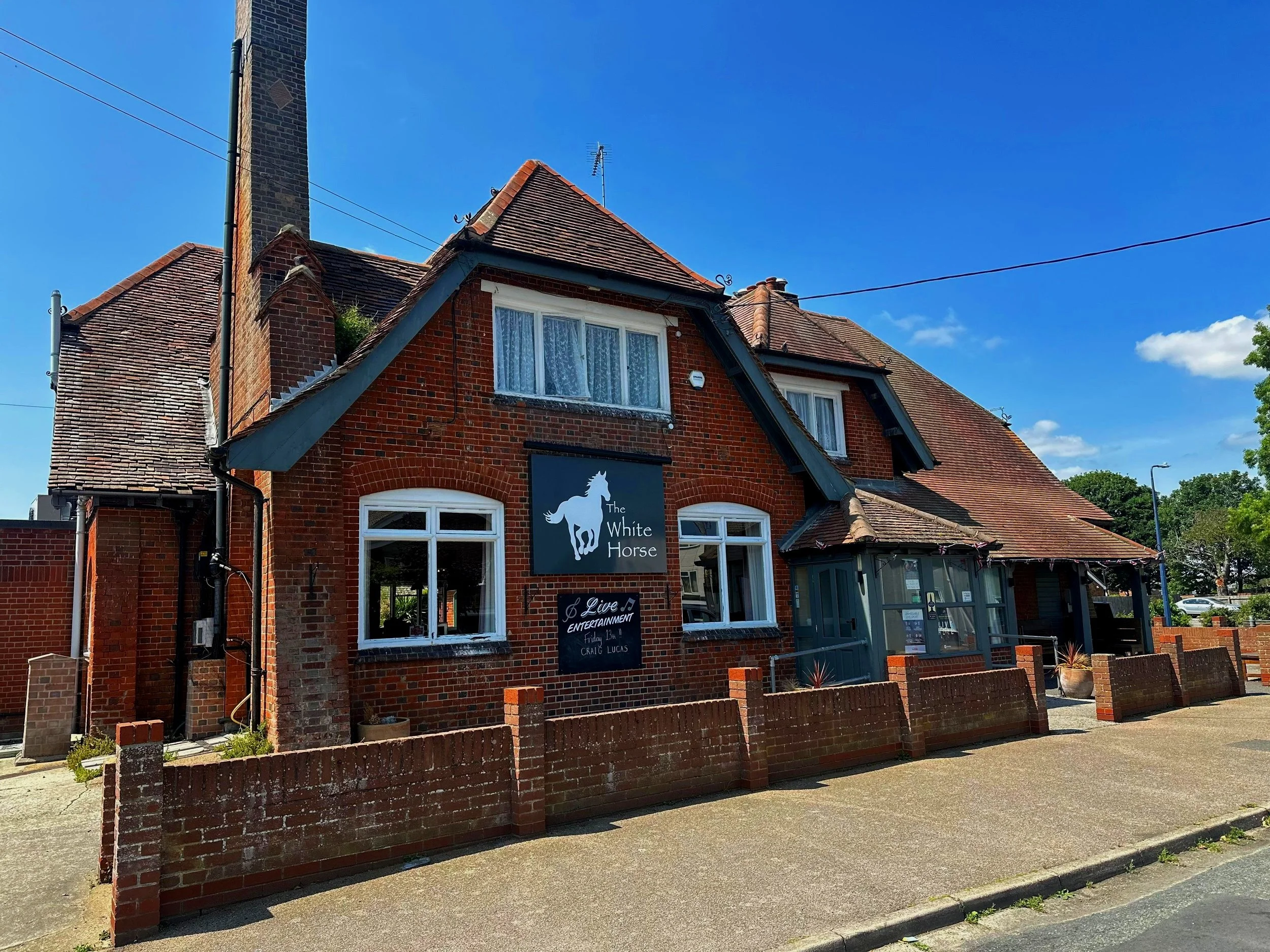 A two-story brick building with a sign for The White Horse pub, featuring a white horse logo, with windows, a small front garden, and a sidewalk under a blue sky.