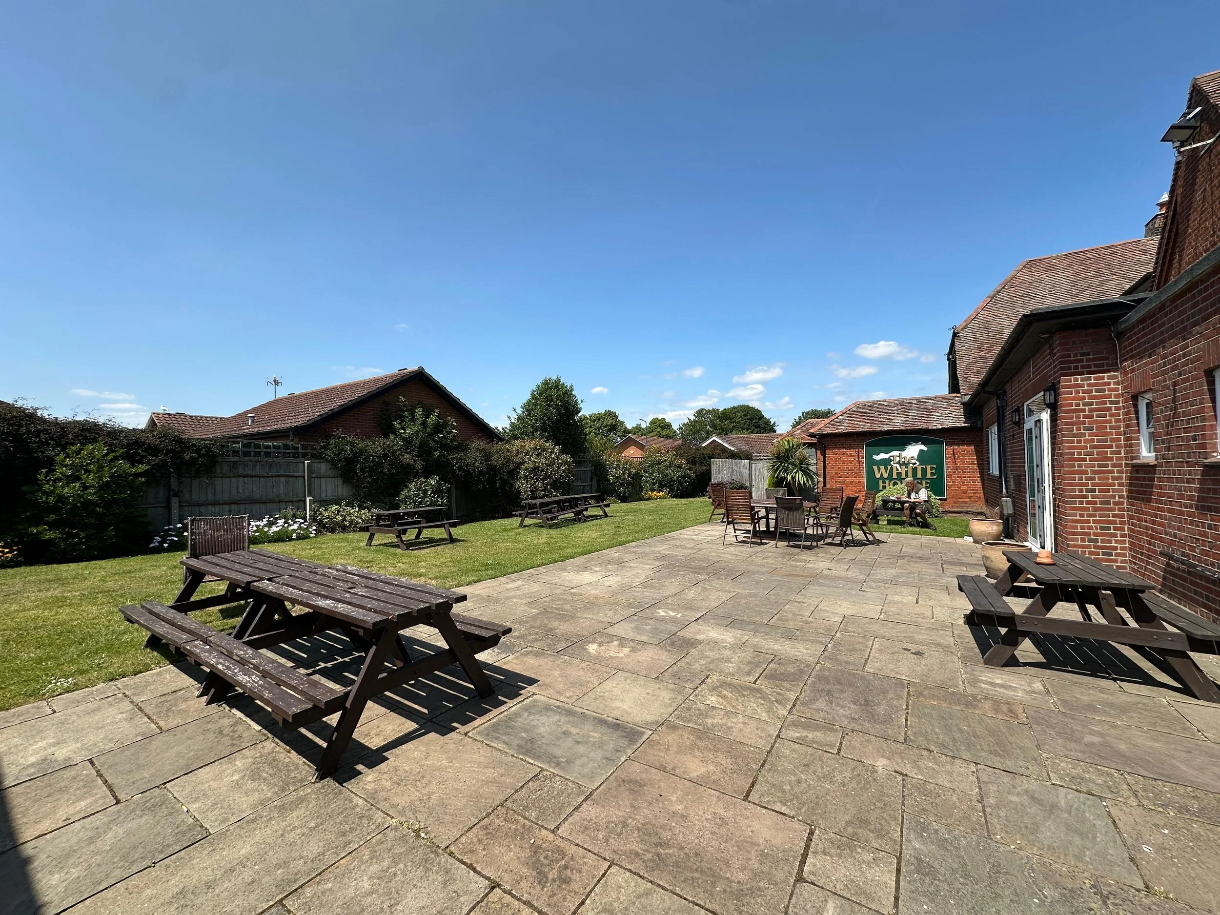 Backyard patio with wooden benches, a dining table with chairs, green grass, bushes, and houses with brick walls under a blue sky.