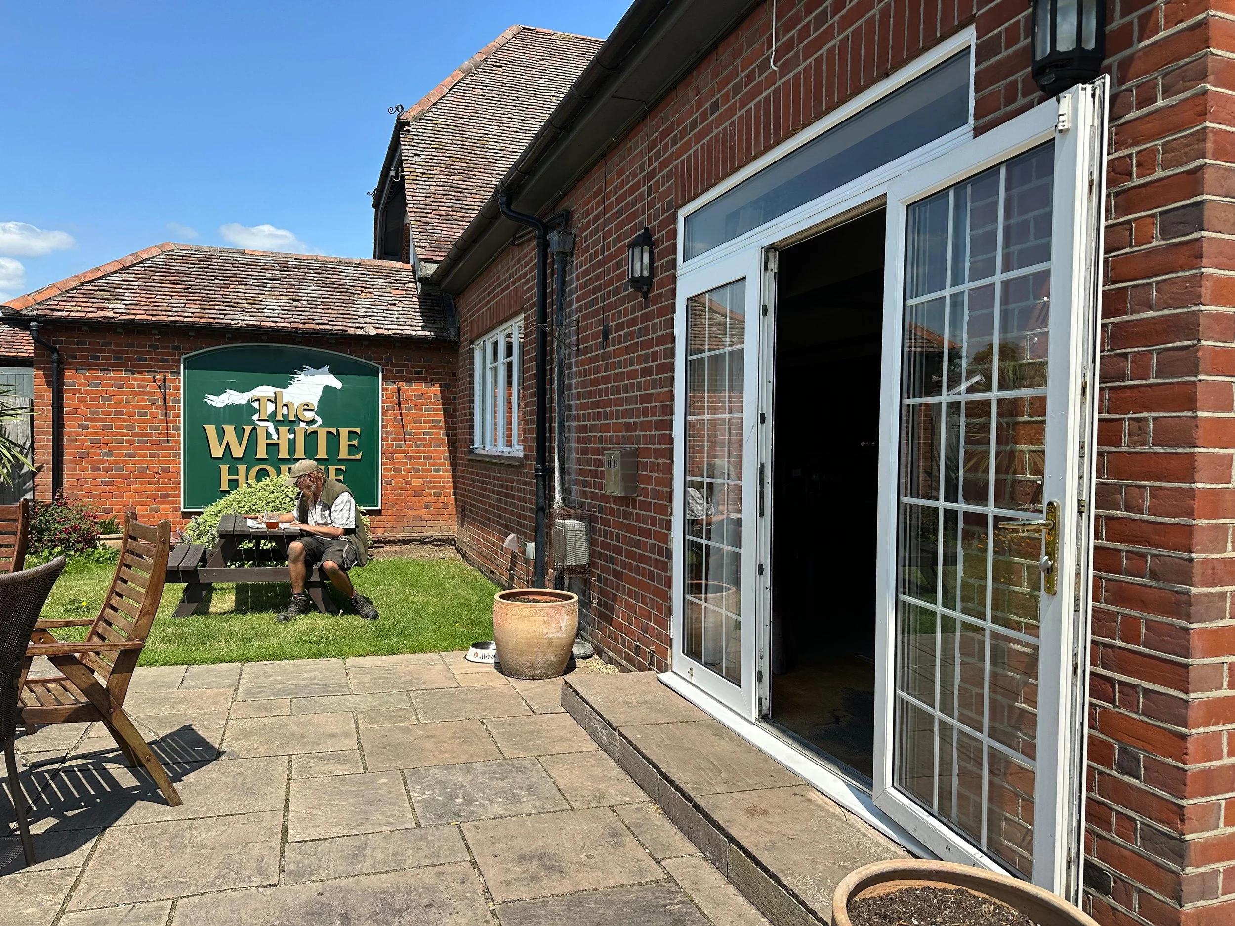 Backyard patio with a man sitting at a picnic table, brick house with glass door, and a green sign for The White Horse pub in the background.